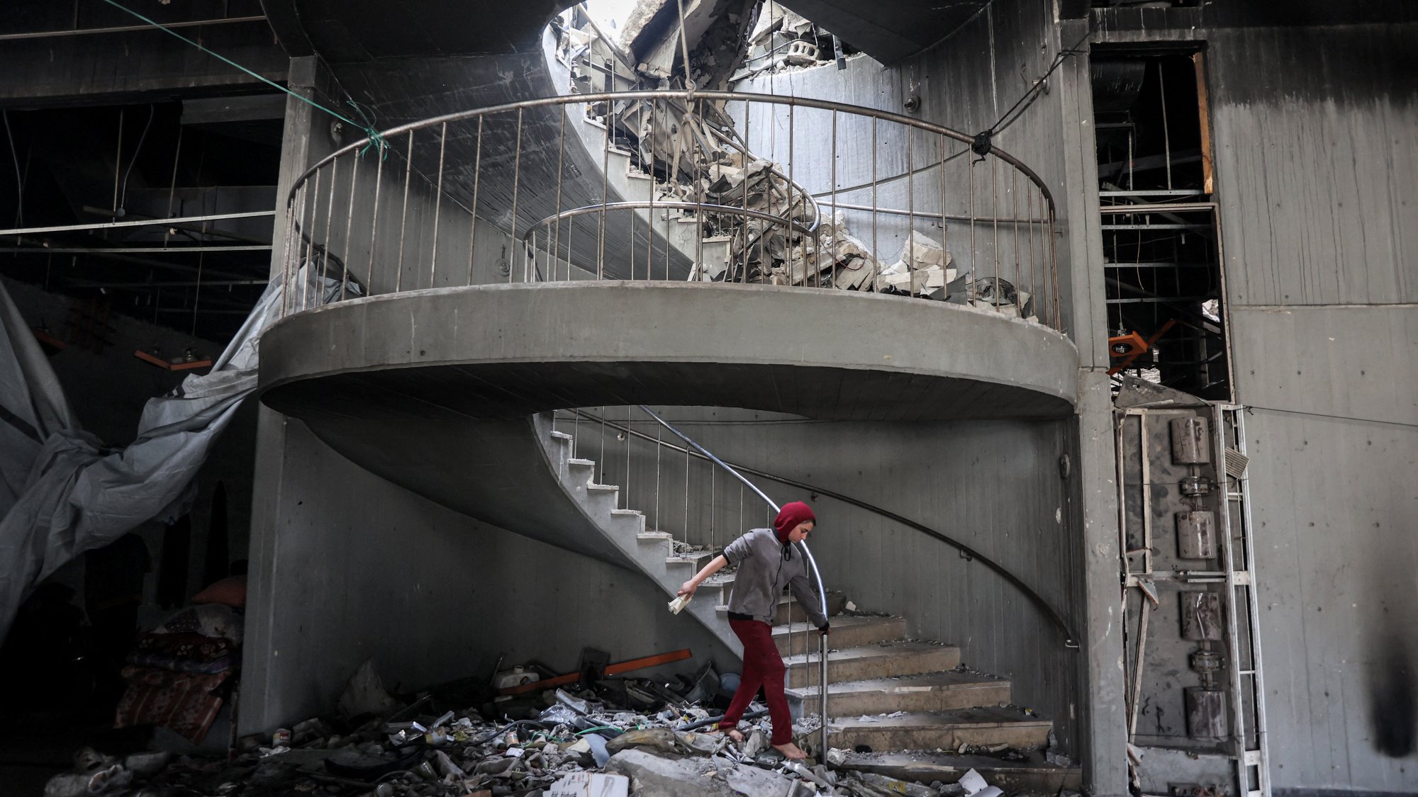 L'image montre un escalier en colimaçon partiellement détruit, avec des débris éparpillés autour. Le plafond a subi d'importants dégâts, laissant apparaître un trou par lequel la lumière pénètre. Un individu, utilisant des béquilles, se déplace prudemment sur les marches. L'ambiance générale évoque des conditions de délabrement et de désolation, probablement à la suite d'un événement catastrophique.