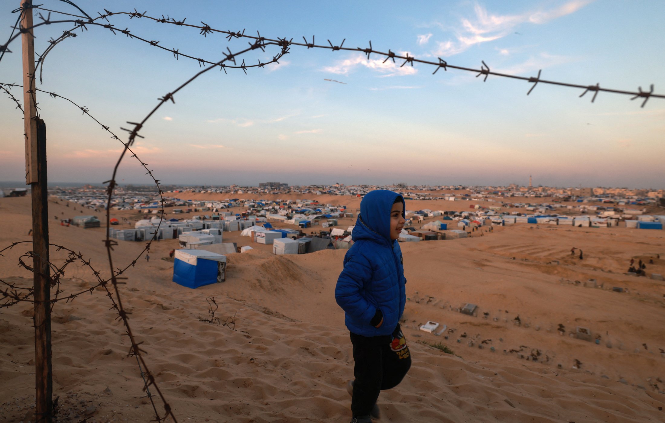 L'image montre un jeune garçon portant une veste bleue qui marche dans un paysage désertique. En arrière-plan, on aperçoit un camp de réfugiés avec de nombreuses tentes et abris disposés sur le sol sablonneux. Un barbelé est visible au premier plan, ajoutant une dimension de sécurité ou de séparation à la scène. Le ciel est partiellement dégagé, avec des teintes de bleu et d'orange, suggérant peut-être la fin de la journée. L'ensemble transmet une atmosphère à la fois de vulnérabilité et d'espoir.