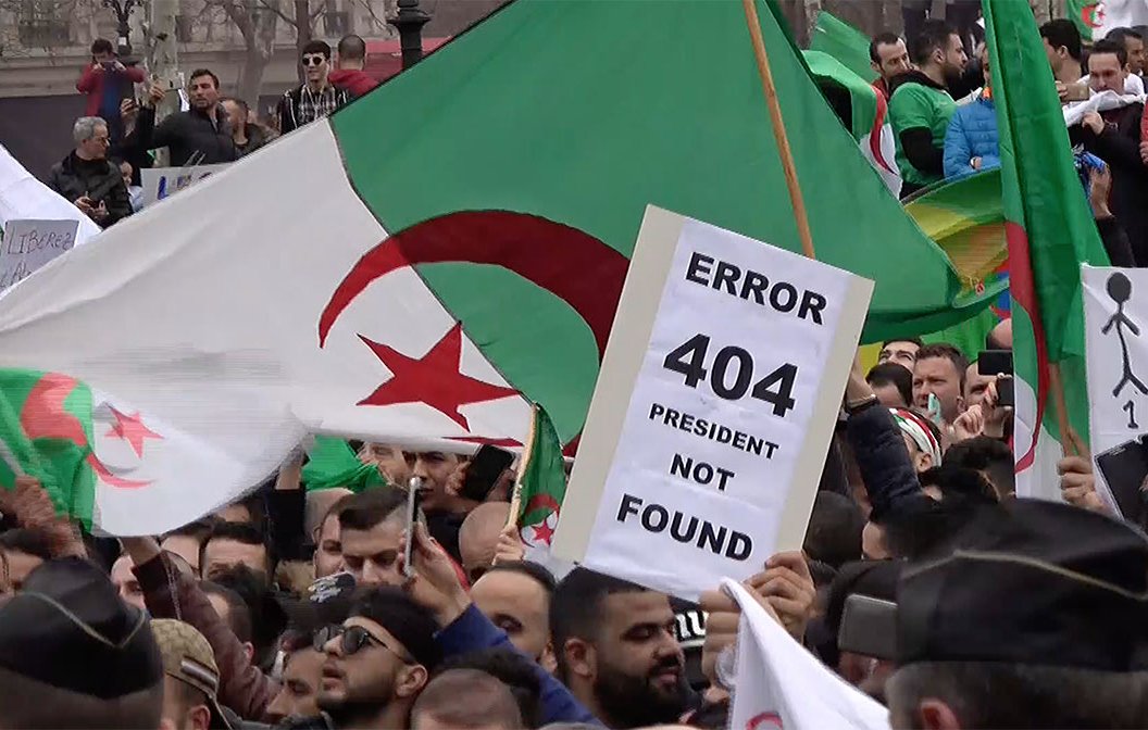 L'image montre une manifestation avec de nombreuses personnes brandissant des drapeaux algériens. Au premier plan, un manifestant tient une pancarte avec l'inscription "ERROR 404 PRESIDENT NOT FOUND", suggérant un mécontentement vis-à-vis de la situation politique. L'ambiance semble être celle d'une contestation, avec une foule réunie pour exprimer ses revendications.
