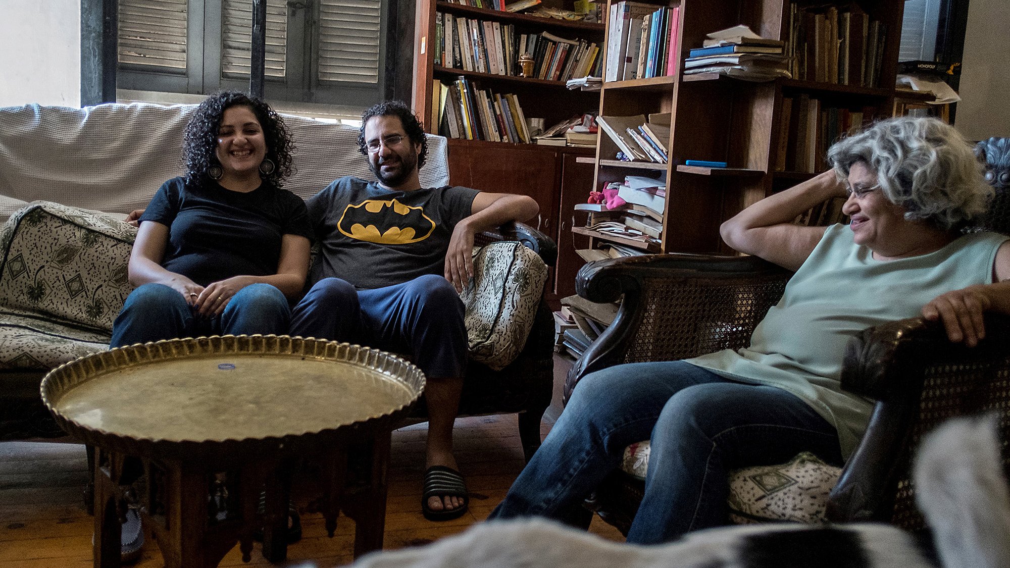 L'image montre un groupe de trois personnes assises dans un salon. Il y a deux femmes et un homme. La première femme, à gauche, sourit et porte un t-shirt noir. L'homme, au centre, porte un t-shirt avec le logo de Batman. La deuxième femme, à droite, a les cheveux bouclés et semble détendue. Ils sont assis sur des canapés, entourés d'étagères remplies de livres et d'objets décoratifs. Une ambiance conviviale et décontractée se dégage de la scène.