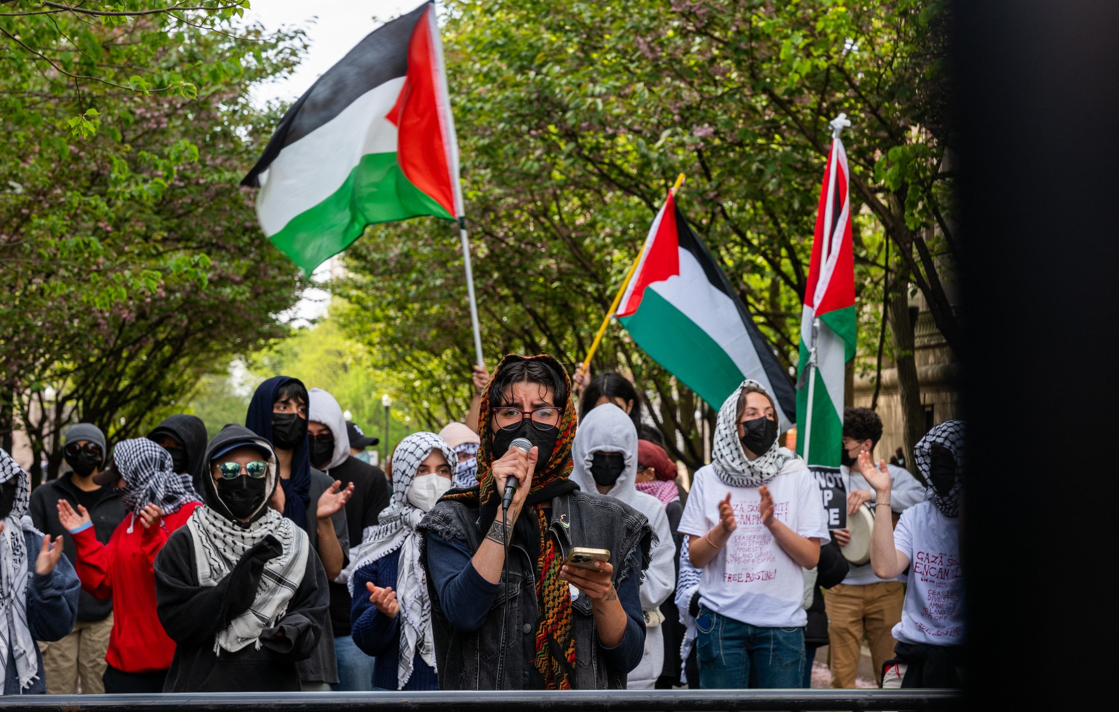 L'image montre une manifestation en plein air où plusieurs personnes sont rassemblées. Au premier plan, une personne prend la parole avec un microphone, vêtue de vêtements sombres et entourée de manifestants portant des masques. Beaucoup des présents arbore des foulards ou keffiehs. En arrière-plan, des drapeaux palestiniens flottent, ajoutant de la couleur à la scène. Les manifestants semblent soutenir une cause, probablement liée à la Palestine, et il y a une atmosphère de solidarité parmi le groupe. Les arbres en fleurs encadrent la scène, créant un contraste avec l'intensité de la manifestation.