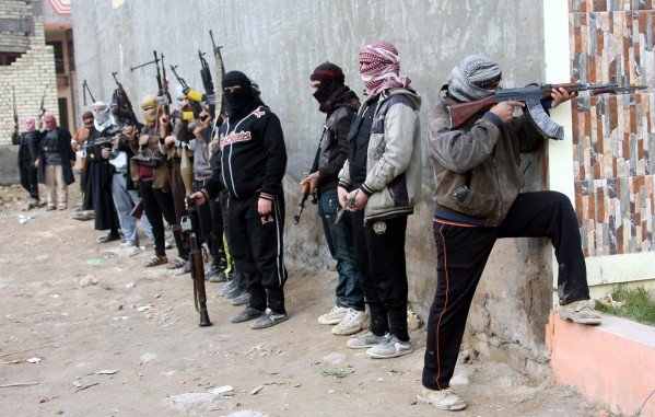 L'image montre un groupe de personnes armées, se tenant alignées le long d'un mur. Elles portent des vêtements sombres et des couvertures sur la tête, ce qui masque leurs visages. Chacune d'elles tient une arme à feu, et certaines adoptent des postures prêtes au combat. L'environnement semble urbain, avec des bâtiments en arrière-plan et un terrain sale. L'atmosphère générale est tendue et évoque un contexte de conflit ou de violence.