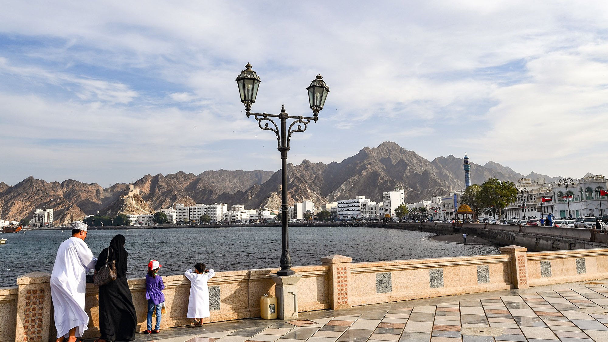 L'image montre une scène de bord de mer, probablement à Muscat, Oman. Dans le premier plan, on peut voir une famille vêtue de vêtements traditionnels. Un homme et une femme se tiennent près d'un lampadaire, regardant vers l'horizon. À côté d'eux, deux enfants, dont un porte une casquette rouge. En arrière-plan, on aperçoit des montagnes majestueuses ainsi que des bâtiments blancs typiques de la région. Le ciel est partiellement nuageux, créant une ambiance paisible et contemplative.