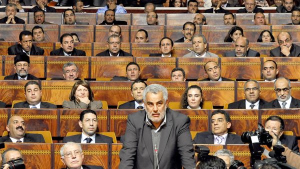 L'image montre une séance dans un bâtiment législatif, où une personne (probablement un membre du gouvernement ou un habile orateur) se tient devant un grand groupe de personnes assises. Les membres de l'assemblée, en arrière-plan, semblent concentrés sur le discours prononcé. On peut voir des journalistes avec des caméras, suggérant que cet événement est couvert par les médias. L'atmosphère est formelle et sérieuse, typique de débats ou de discussions politiques.