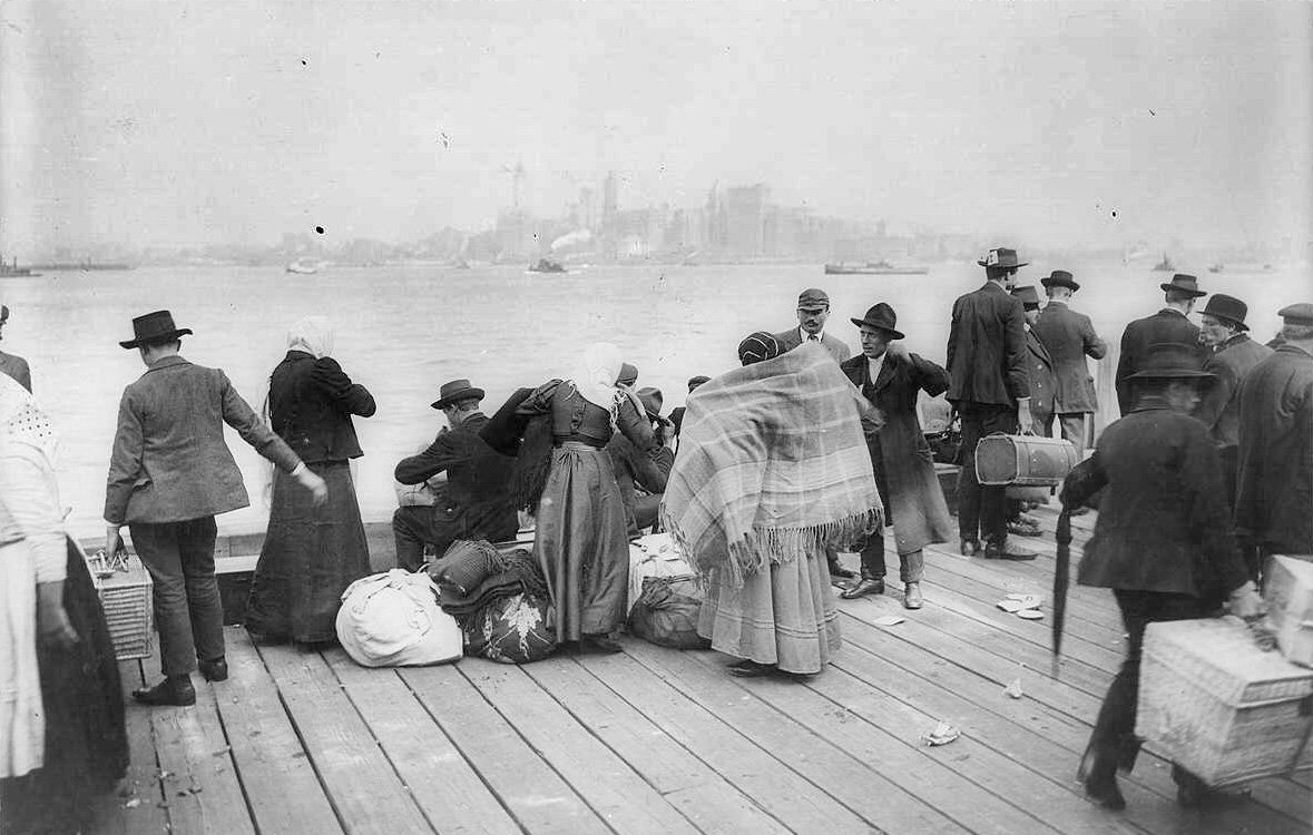 L'image montre un groupe de personnes rassemblées sur un quai, probablement à une époque antérieure. On peut voir des individus portant des habits typiques de l'époque, dont des chapeaux, et certains sont emmitouflés dans des couvertures. Ils semblent s'affairer autour de bagages et de marchandises, évoquant des thèmes de migration ou de transit. En arrière-plan, on aperçoit des bâtiments ainsi que des bateaux sur l'eau, suggérant un port animé. L'atmosphère semble à la fois chargée et historique.