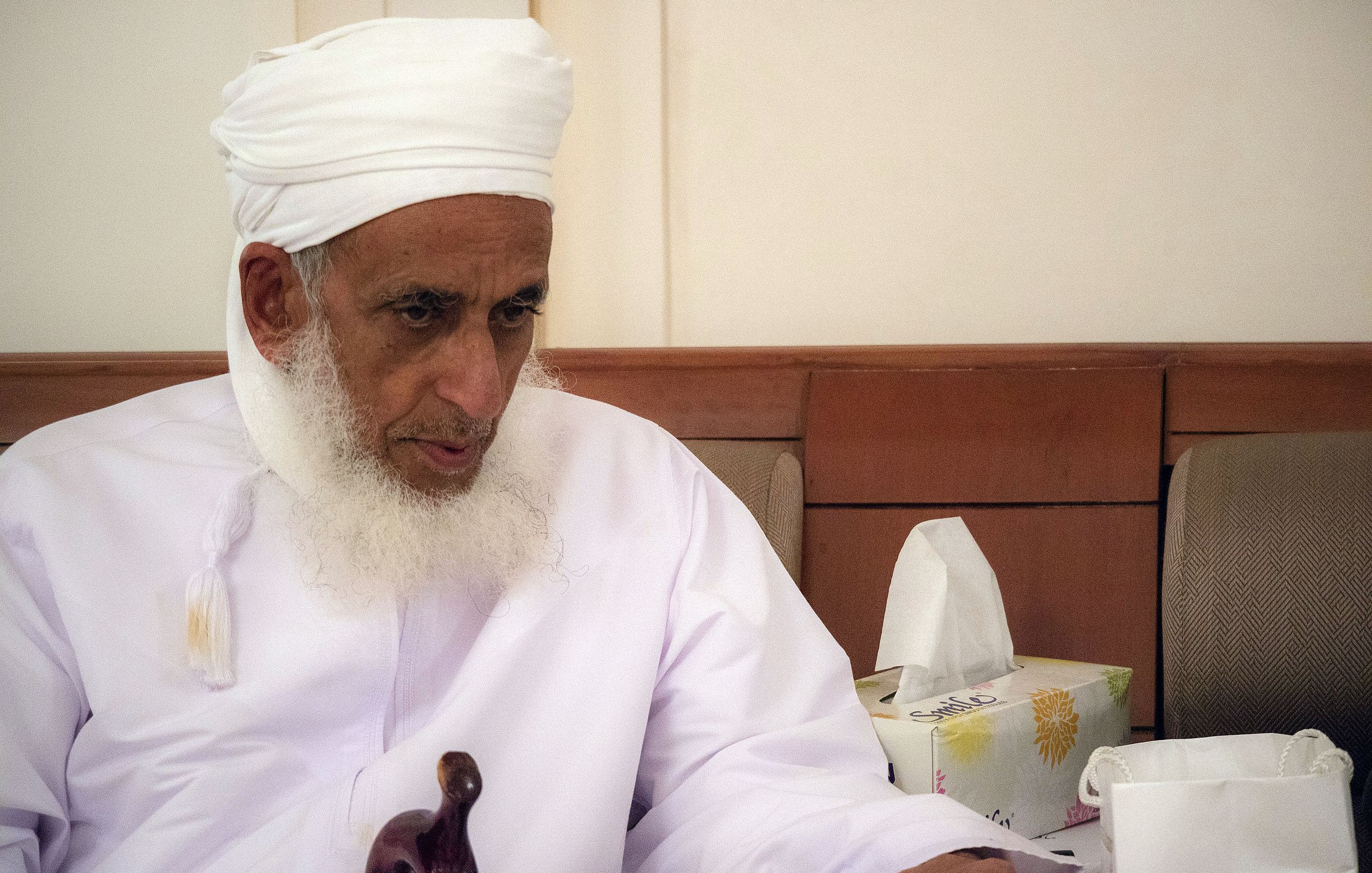 L'image montre un homme âgé portant un turban blanc et une robe traditionnelle. Il a une barbe grise et semble pensif, assis sur un canapé. À côté de lui, on peut voir des mouchoirs et un sac en papier, ce qui suggère qu'il se trouve dans un environnement intérieur calme. L'éclairage est doux, créant une atmosphère sereine.