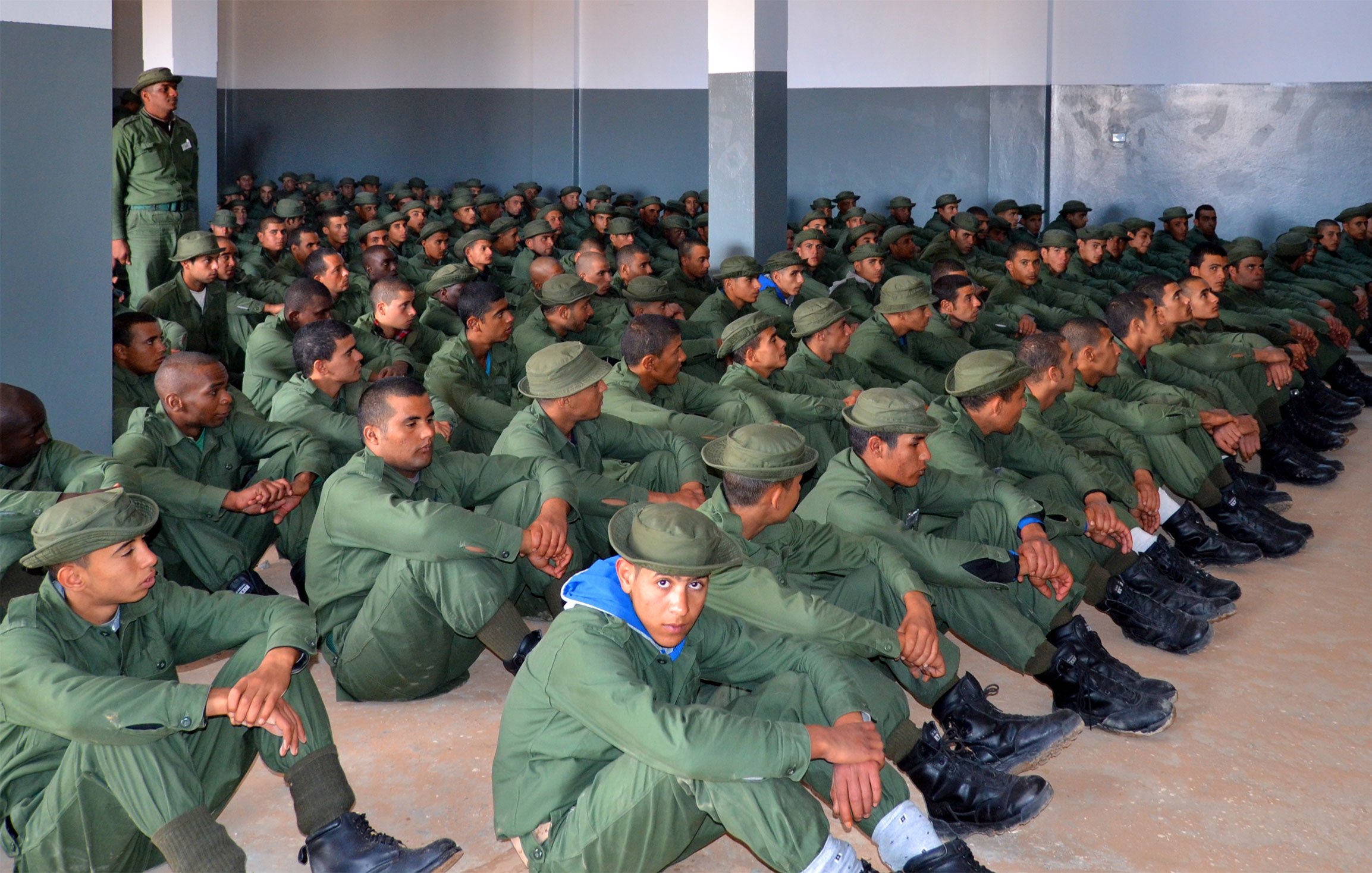 L'image montre un grand groupe de soldats assis dans une salle. Ils portent des uniformes verts et des bottes noires, et sont alignés en rangées sur le sol. Un instructeur, également en uniforme, se tient debout devant eux, semblant donner des instructions ou une formation. Les murs de la salle sont peints en gris, et l'atmosphère semble sérieuse et concentrée.