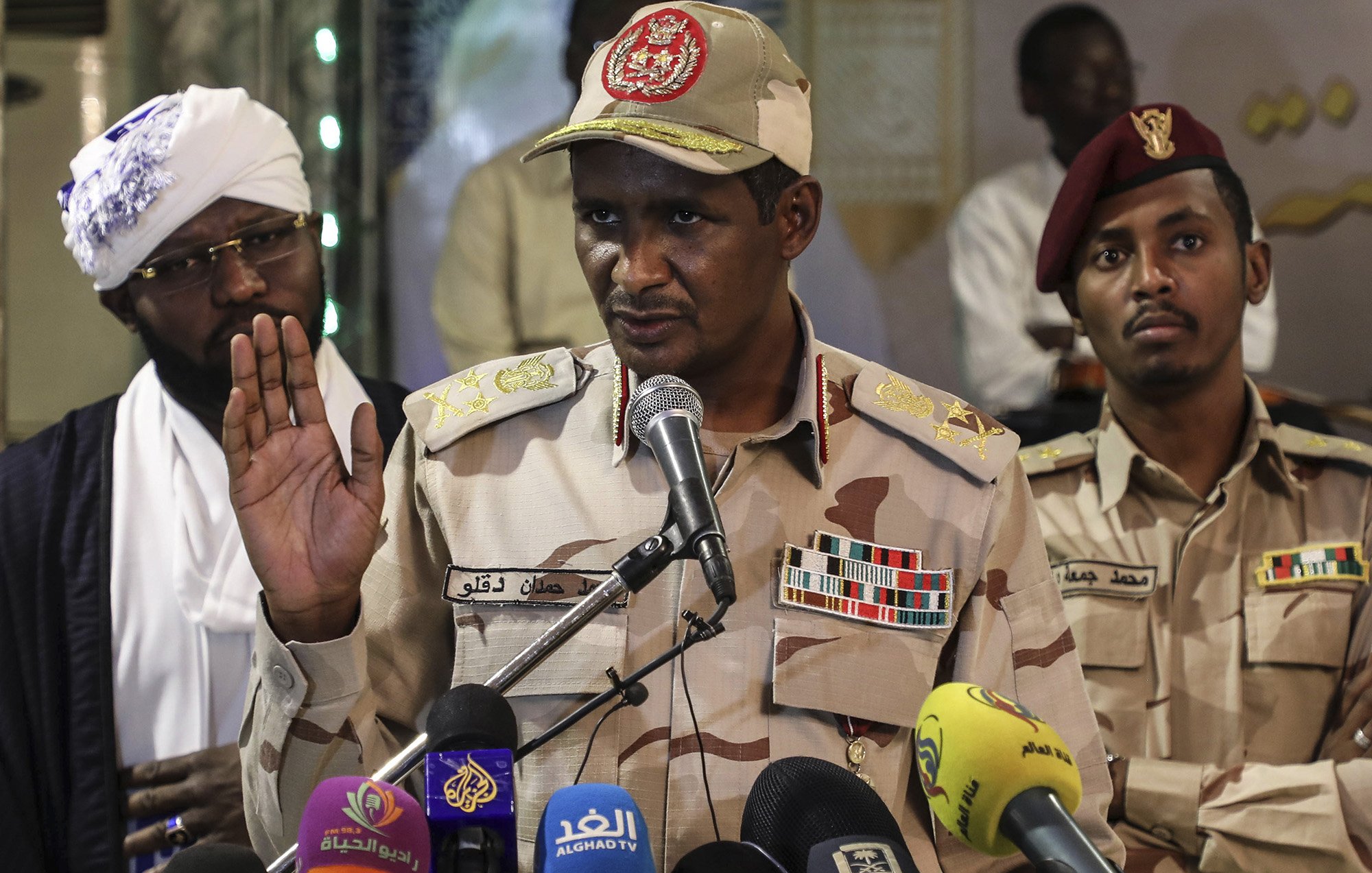 L'image montre un homme en uniforme militaire, portant une casquette décorée, qui s'exprime devant un micro lors d'une conférence de presse. Il semble être en train de faire une déclaration importante, avec une main levée. En arrière-plan, deux autres hommes sont également présents, l'un portant un chapeau traditionnel et l'autre en uniforme militaire. Divers microphones de médias sont placés devant lui. L'ambiance semble sérieuse, suggérant un contexte politique ou militaire.