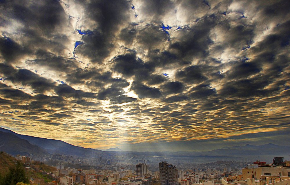 Cette image montre un paysage urbain avec des bâtiments s'étendant dans la vallée, sous un ciel dramatique rempli de nuages. Les nuages sont denses et variés, créant une texture visuelle intéressante, tandis que des rayons de lumière percent à travers eux, illuminant partiellement la ville en contrebas. On peut également apercevoir des montagnes à l'arrière-plan, ajoutant une dimension naturelle à la scène. L'atmosphère est à la fois majestueuse et sereine, capturant un moment poétique de la journée.
