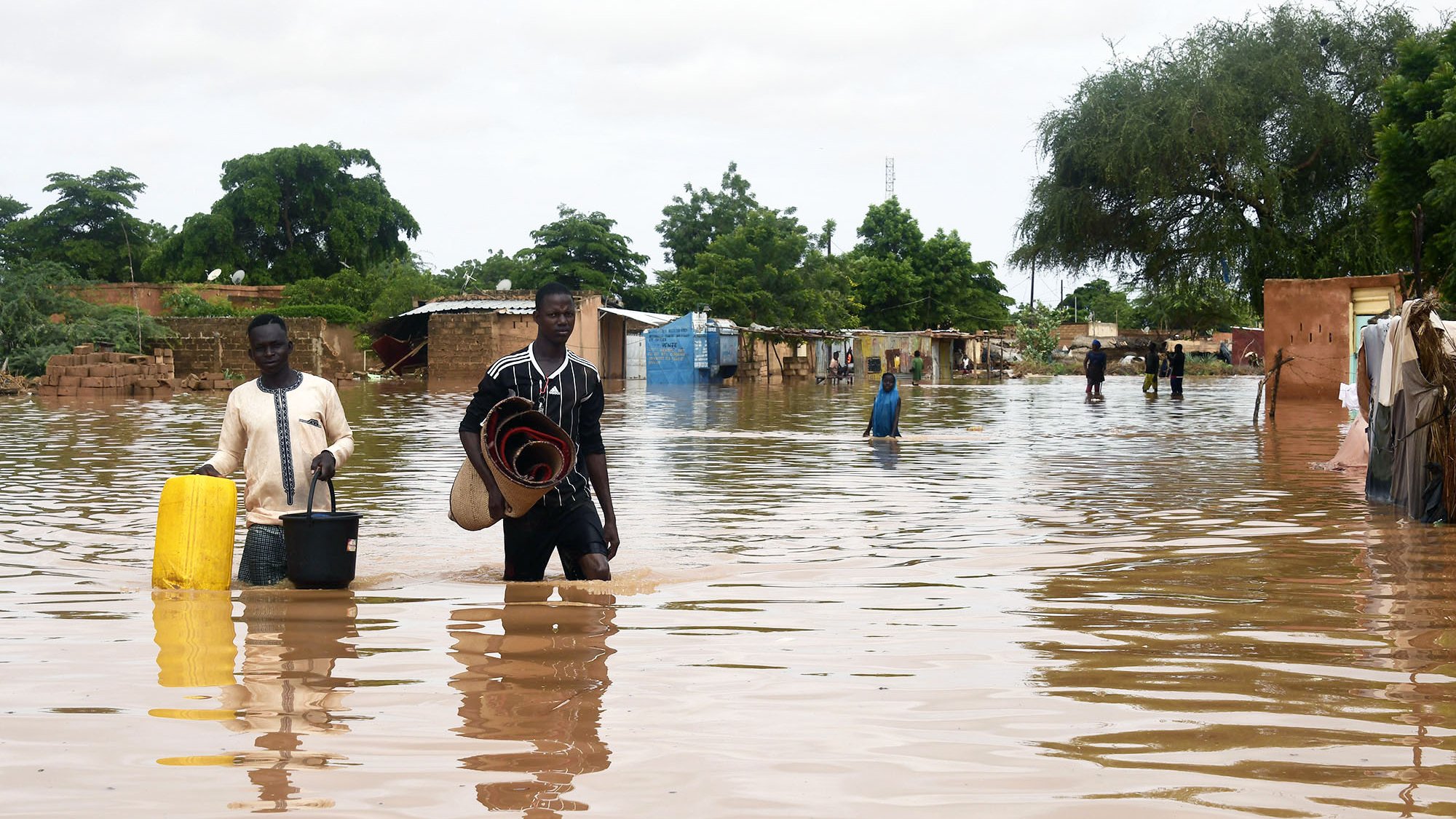 L'image montre une scène de inondation où deux hommes marchent dans une zone inondée. L'eau monte jusqu'à leurs jambes et environ une partie des maisons environnantes est submergée. Les hommes portent des objets, l'un avec un seau et l'autre avec des paniers ou des matelas. En arrière-plan, on peut voir des arbres et des bâtiments partiellement immergés, représentant une situation difficile due aux inondations. Le ciel est nuageux, ce qui suggère des conditions météorologiques instables.