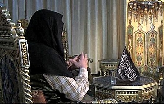 L'image montre deux personnes assises dans un cadre décoratif. L'un des individus porte un hijab et est assis sur un fauteuil orné, tandis que l'autre, en costume, est également assis sur un fauteuil face à lui, tenant un livre. L'environnement semble richement décoré avec des motifs orientaux et des lumières suspendues. L'atmosphère de la scène semble être celle d'une conversation sérieuse.