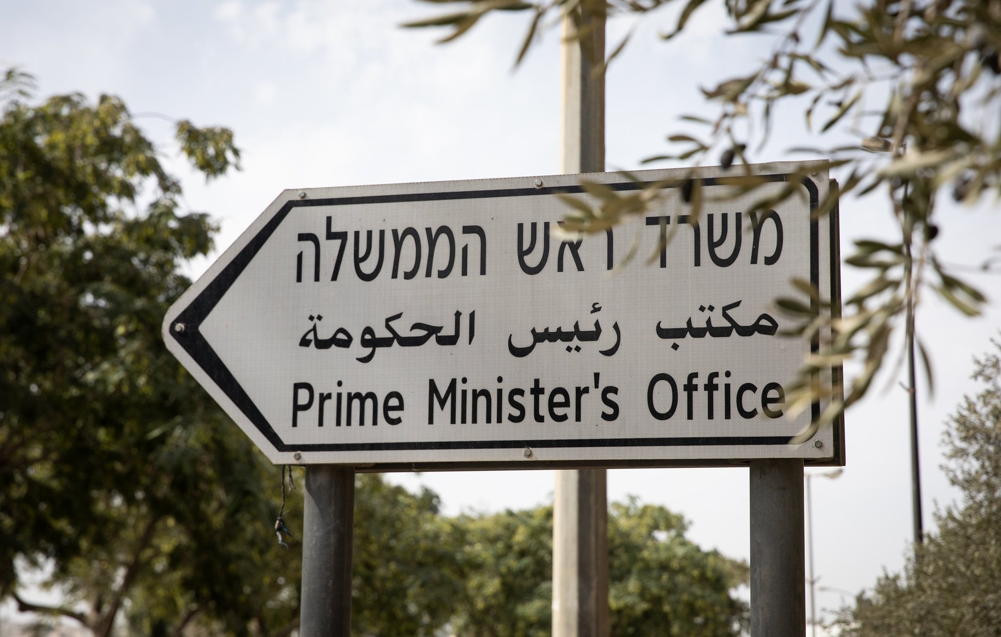 L'image montre un panneau de signalisation indiquant le "Bureau du Premier Ministre". Ce panneau est trilingue, avec des inscriptions en hébreu, en arabe et en anglais, ce qui reflète la diversité linguistique de la région. Le panneau est installé dans un environnement avec des arbres en arrière-plan.