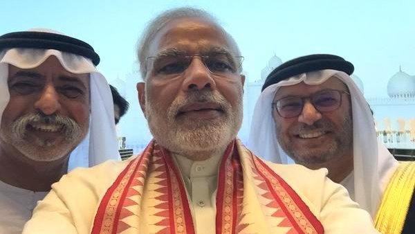 L'image montre un groupe de trois hommes prenant un selfie, probablement dans un cadre officiel ou lors d'un événement. L'homme au centre porte un vêtement traditionnel avec un foulard coloré autour du cou. Les deux autres hommes à côté de lui portent des vêtements traditionnels du Moyen-Orient, incluant un keffieh et un costume, ajoutant à l'atmosphère culturelle de la scène. En arrière-plan, on aperçoit des bâtiments qui semblent être emblématiques, bien que les détails ne soient pas clairs. L'expression des visages est souriante et conviviale, suggérant une atmosphère amicale.