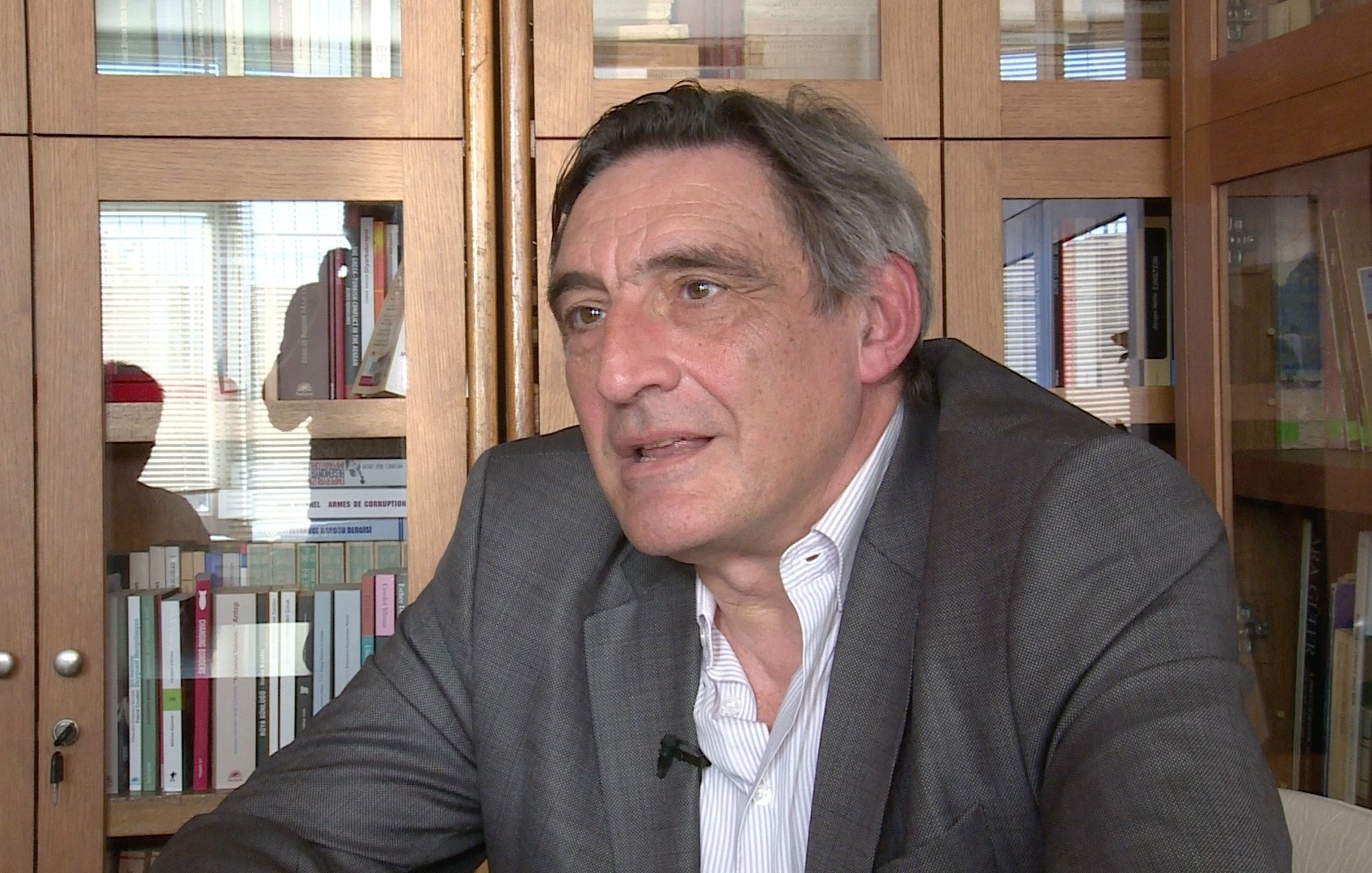 L'image montre un homme assis à une table, vêtu d'un costume gris. Il semble en train de parler ou de réfléchir, avec une expression sérieuse. En arrière-plan, on peut voir des étagères pleines de livres et des objets divers. La lumière naturelle entre par une fenêtre, créant un ambiance chaleureuse dans la pièce.