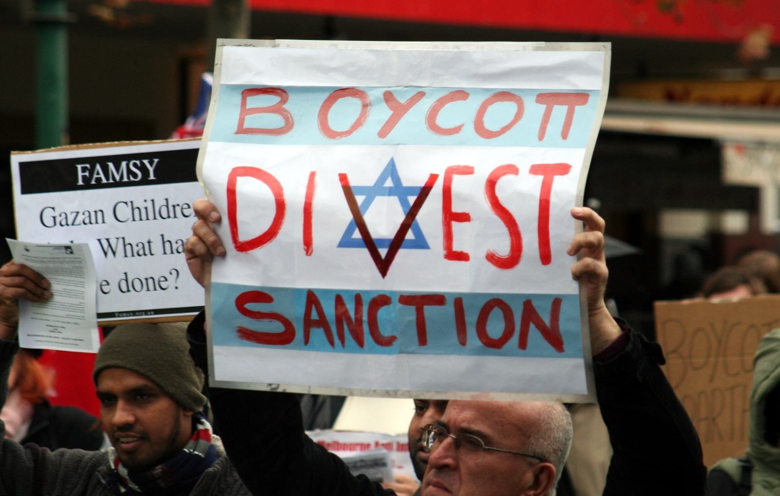 L'image montre des manifestants tenant des pancartes lors d'une manifestation. L'une des pancartes, bien visible, porte les mots "BOYCOTT, DIVEST, SANCTION" avec un symbole en forme d'étoile. Les manifestants semblent exprimés des opinions politiques ou sociales fortes, et l'ambiance de l'événement semble engagée.