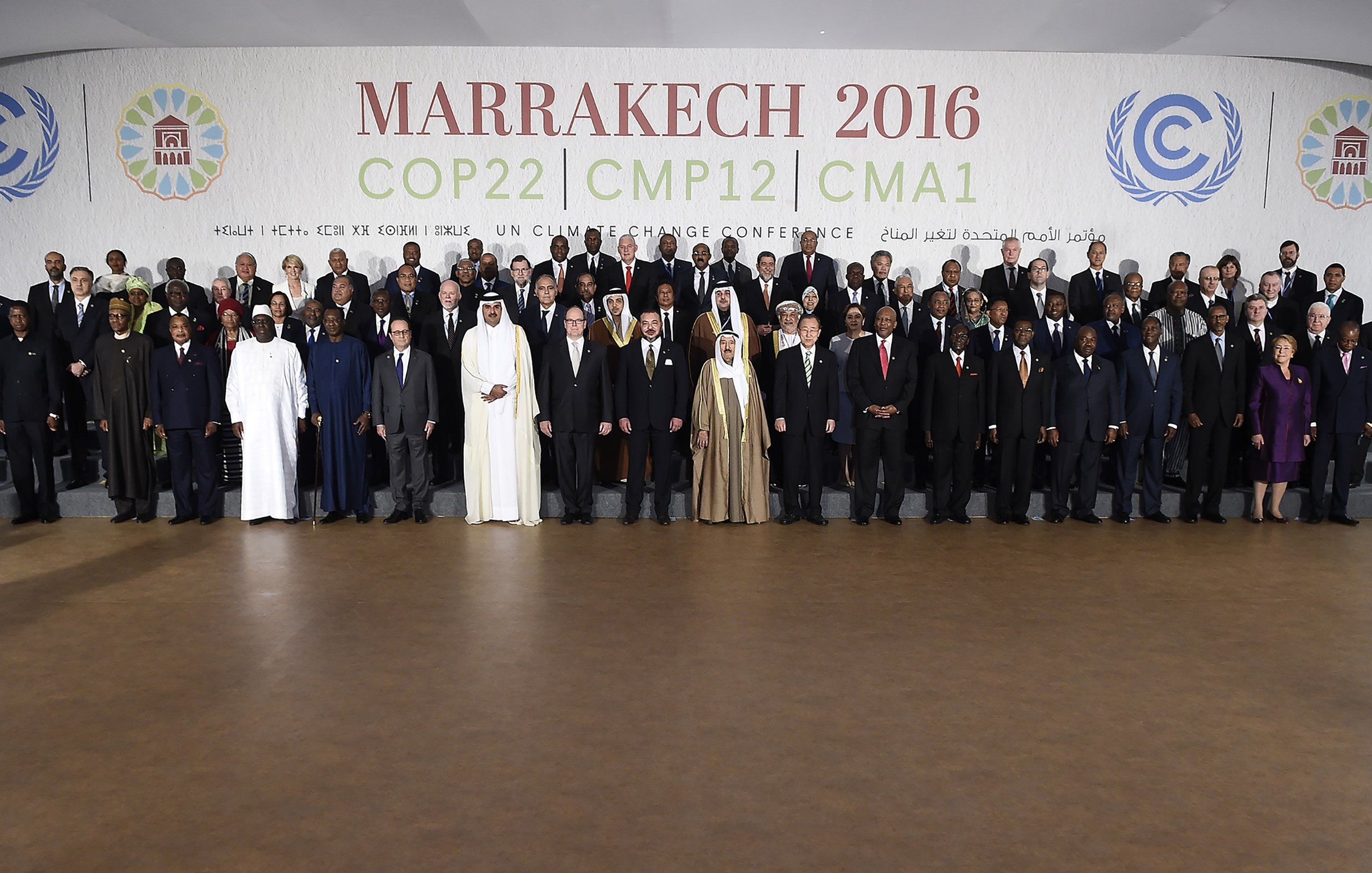 L'image montre un groupe de participants rassemblés lors de la conférence COP22 qui s'est tenue à Marrakech en 2016. Les membres représentés incluent des dirigeants et des délégués de divers pays, vêtus de costumes formels et de tenues traditionnelles. Ils se tiennent debout sur des gradins, souriant pour la photo officielle, avec un fond affichant le nom de l'événement et des logos des Nations Unies. L'ambiance évoque un sentiment de collaboration internationale pour faire face aux défis climatiques.