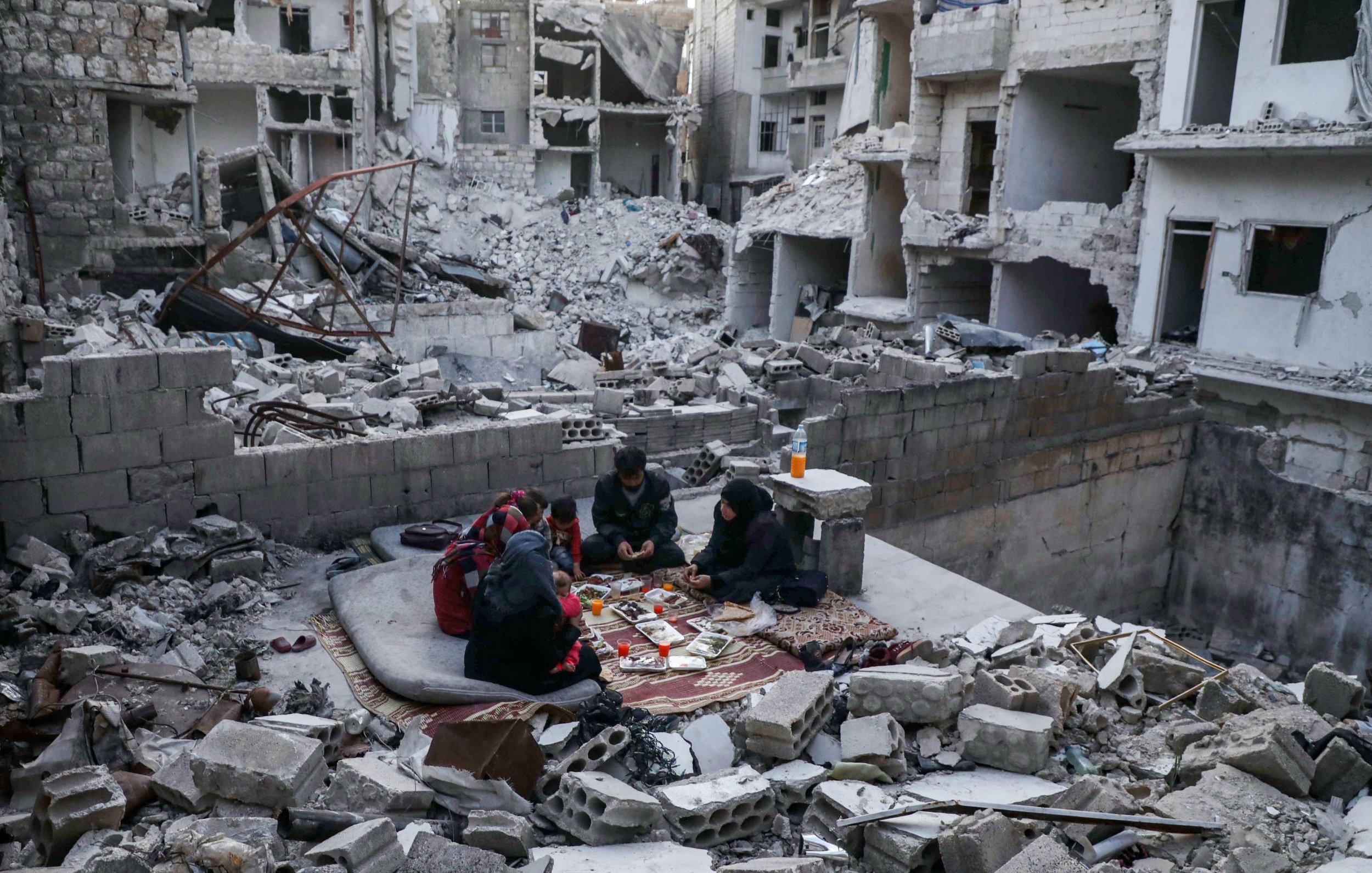L'image montre un groupe de personnes assises sur un tapis dans un environnement dévasté. Autour d'eux, on peut voir des débris de bâtiments effondrés et des structures endommagées. Les personnes semblent concentrées sur une activité, malgré la destruction qui les entoure. L'atmosphère évoque la résilience et la vie qui continue malgré les difficultés.