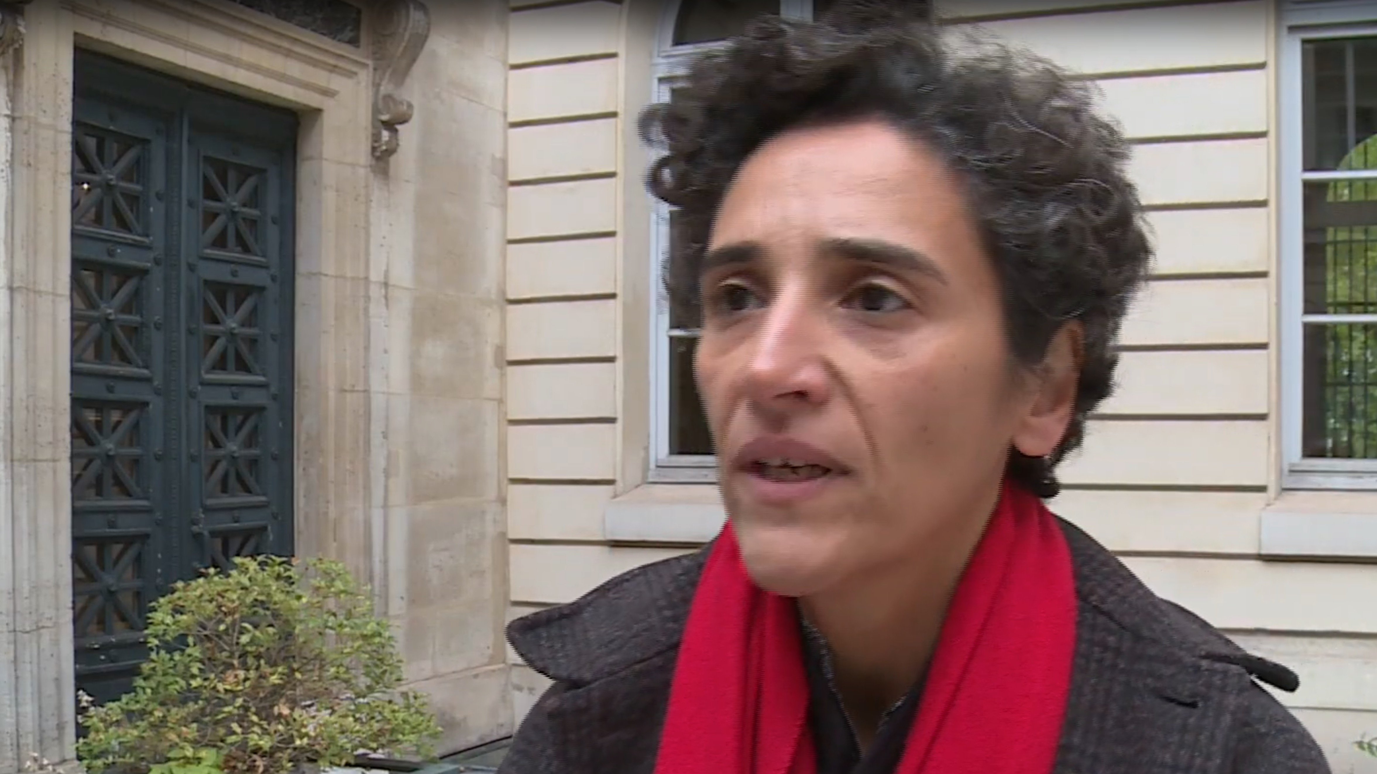 L'image montre une femme avec des cheveux bouclés, portant un manteau sombre et une écharpe rouge. Elle semble s'exprimer avec sérieux, peut-être dans le cadre d'une interview. En arrière-plan, on aperçoit un bâtiment avec des fenêtres et une porte d'entrée, ce qui donne une impression urbaine et architecturale.