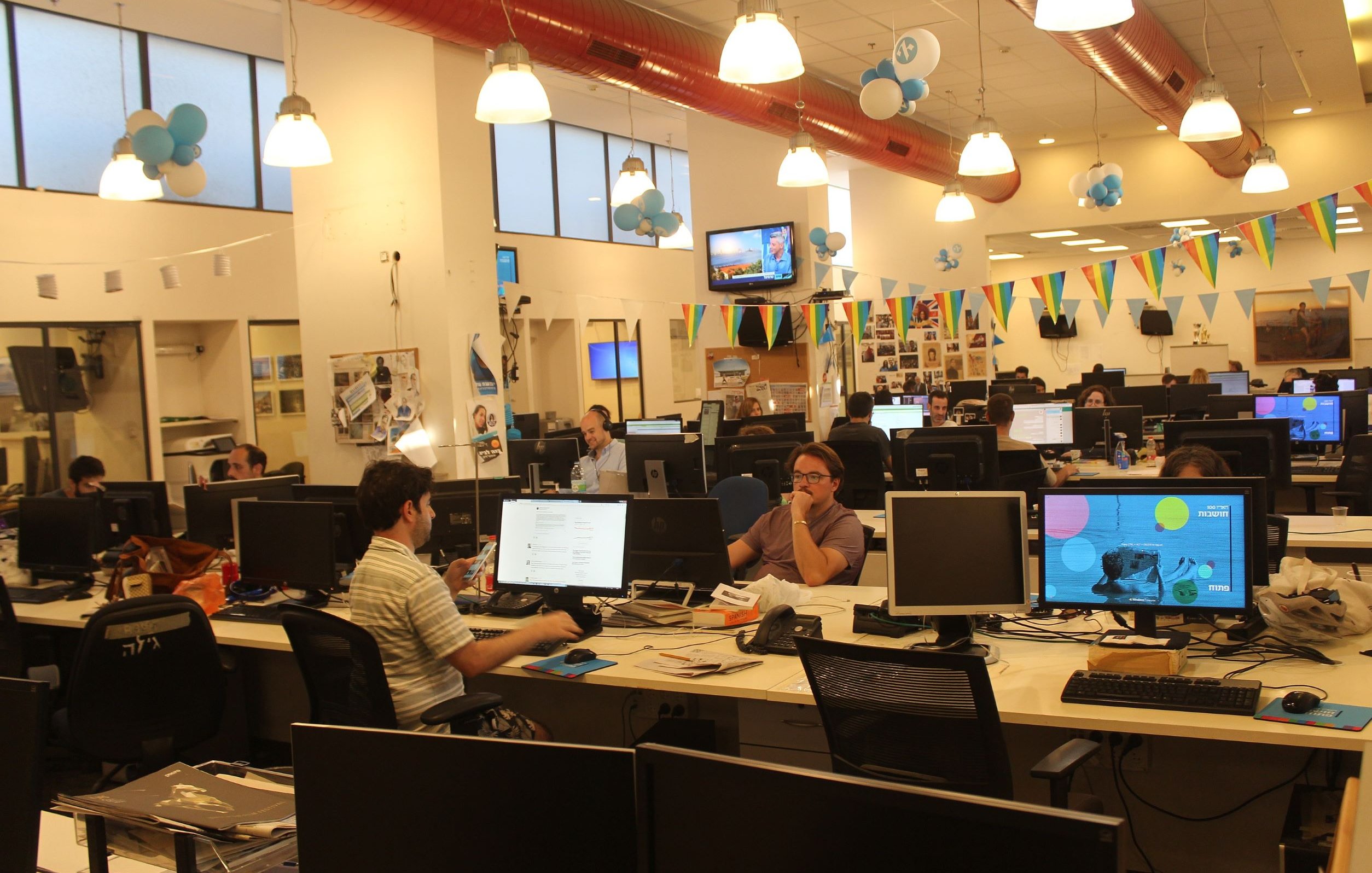 L'image montre un environnement de bureau moderne et animé. Plusieurs employés sont assis devant des ordinateurs, travaillant sur leurs tâches. Les bureaux sont équipés de plusieurs écrans d'ordinateur. La pièce est décorée avec des ballons et des banderoles, suggérant une atmosphère festive ou un événement spécial. La lumière naturelle entre par de grandes fenêtres, ajoutant une ambiance agréable à l'espace de travail.
