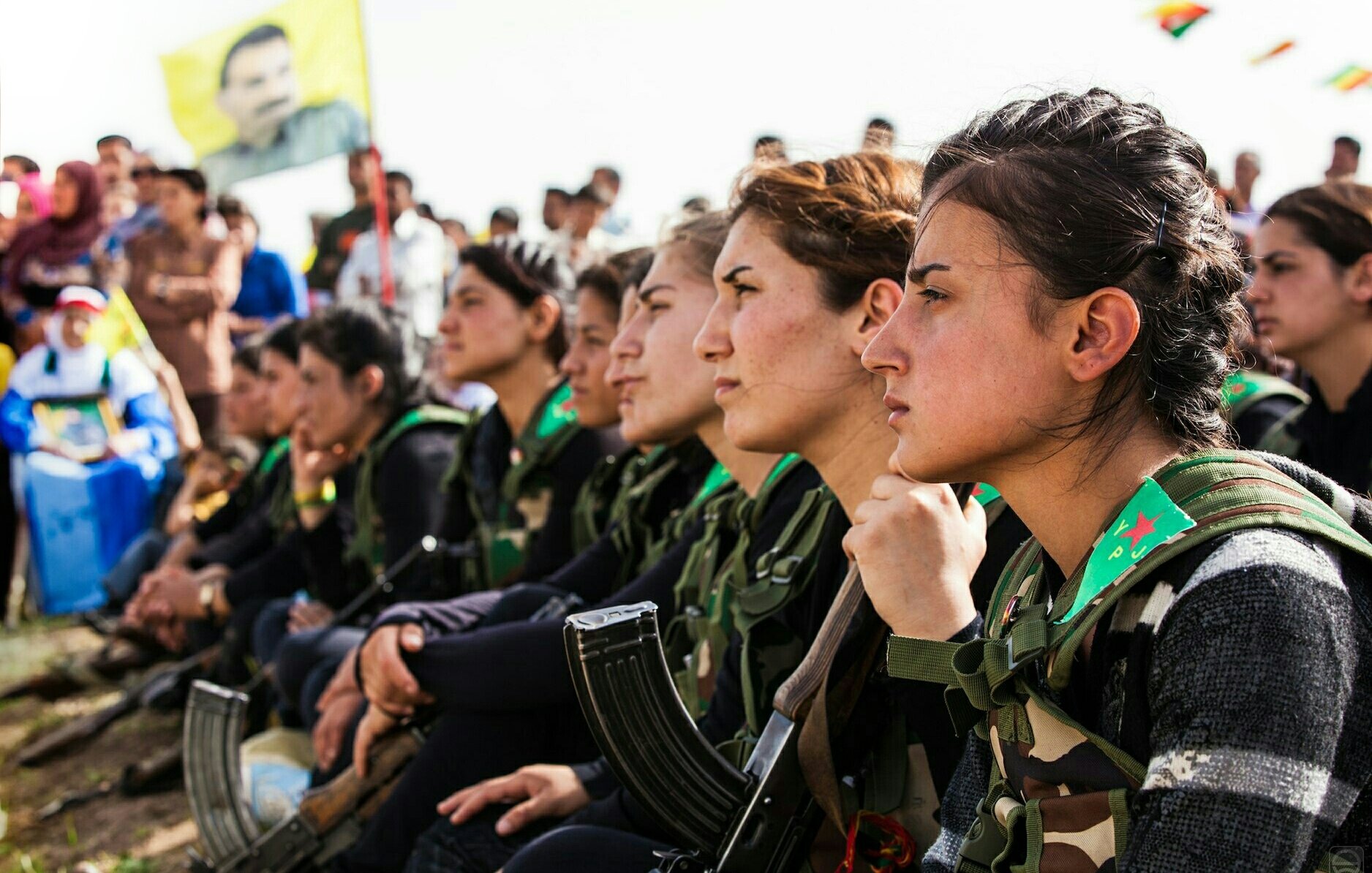 L'image montre un groupe de femmes assises en rang, portant des uniformes militaires avec des armes à leurs côtés. Elles semblent attentives et réfléchies, regardant vers l'avant. En arrière-plan, on peut apercevoir d'autres personnes réunies, ainsi qu'un drapeau symbolique. L'ambiance semble être celle d'un rassemblement ou d'une cérémonie.