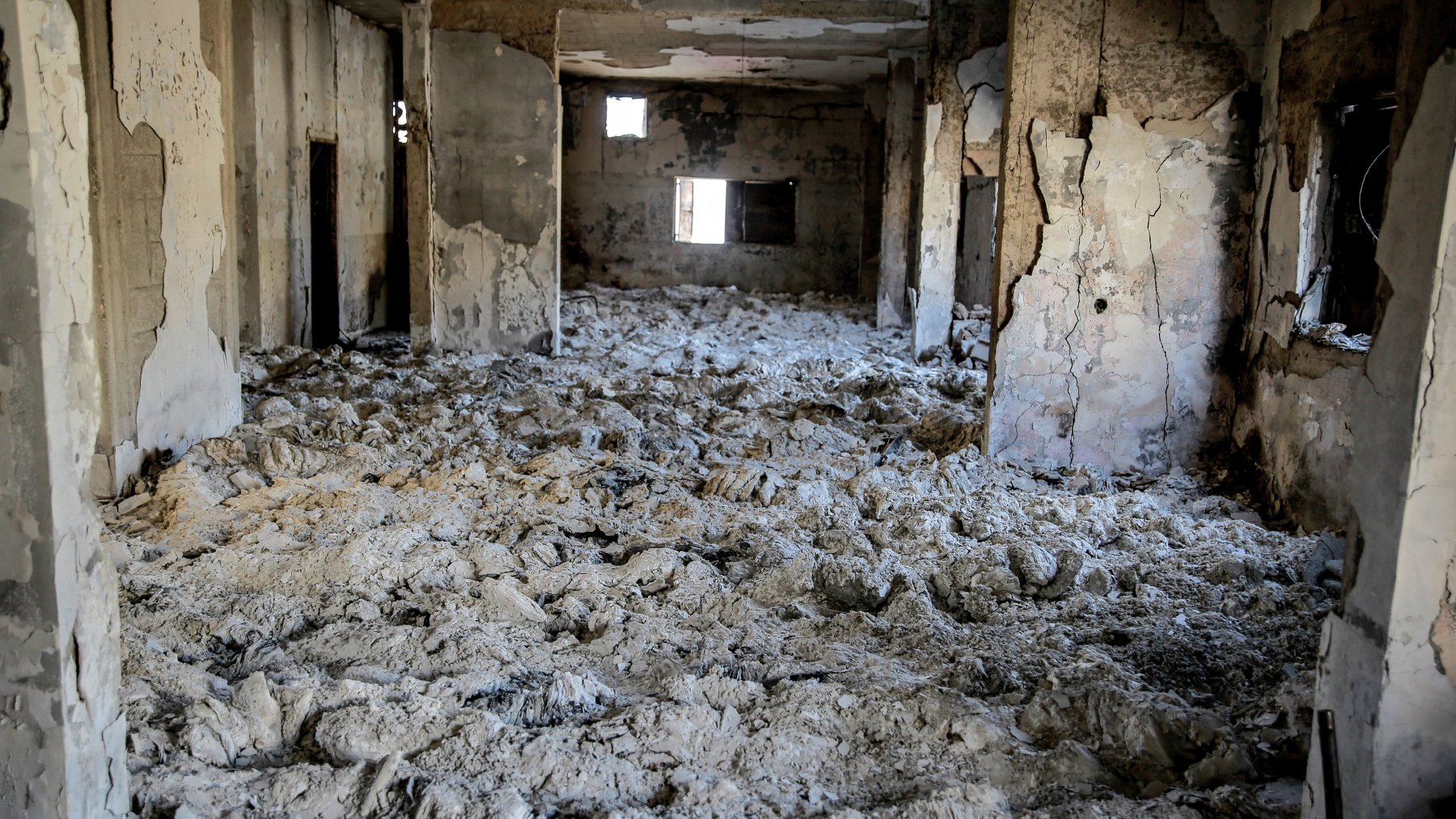 L'image montre l'intérieur d'un bâtiment en ruine, où les murs sont endommagés et décrépis. Le sol est recouvert d'un matériau blanc poussiéreux, probablement des débris ou des restes d'anciennes structures. Les fenêtres sont brisées ou manquantes, créant une atmosphère de désolation et d'abandon. La lumière pénètre à travers les ouvertures, mais l'espace parait sombre et sinistre.