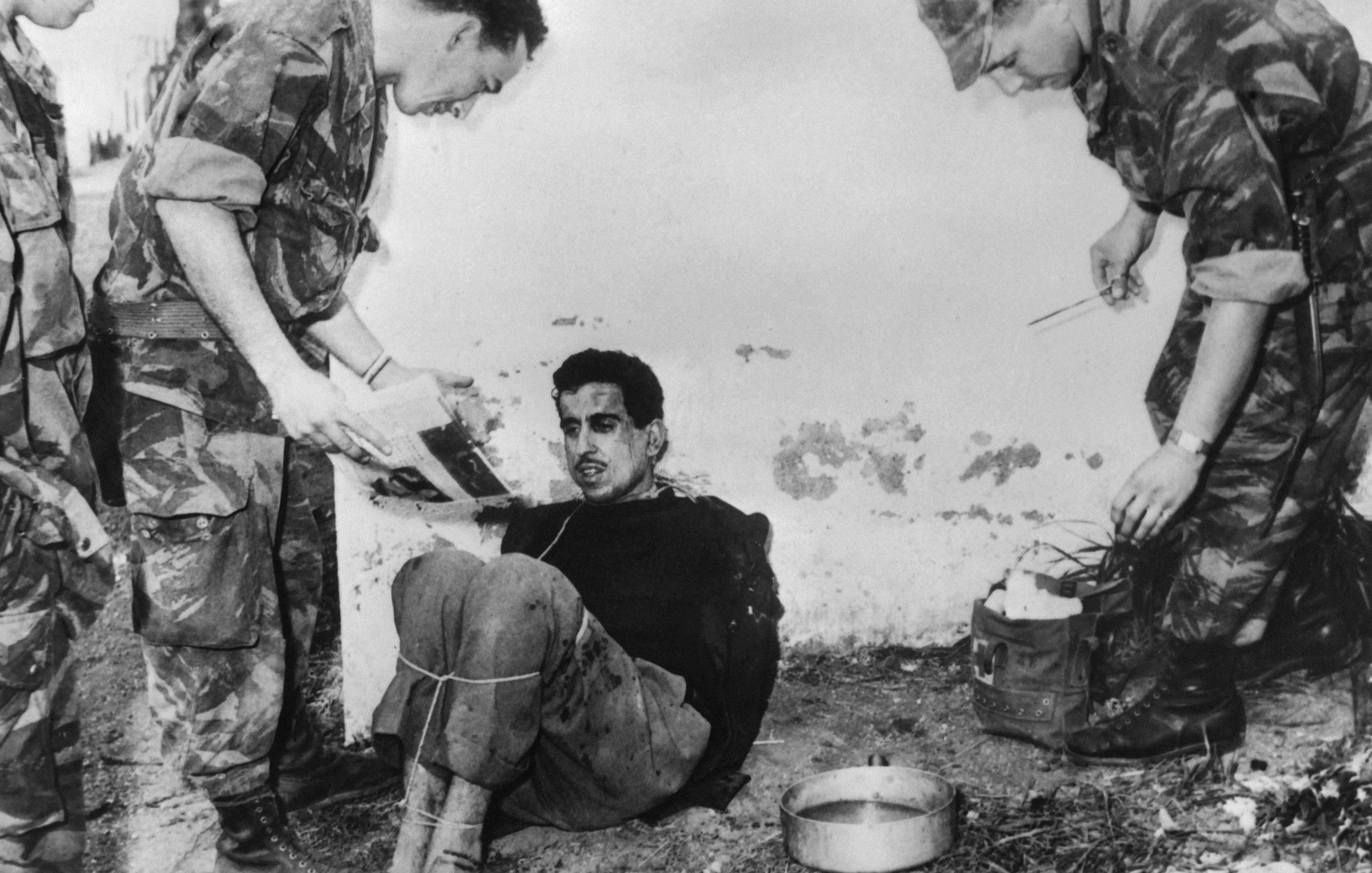L'image montre un homme assis et attaché, visiblement en détresse, tandis que deux soldats, habillés en uniforme militaire, se tiennent près de lui. Un des soldats semble examiner un document ou une photographie, tandis que l'autre s'approche avec un équipement. L'environnement est sobre, et l'ambiance générale de la scène dégage un sentiment de tension.