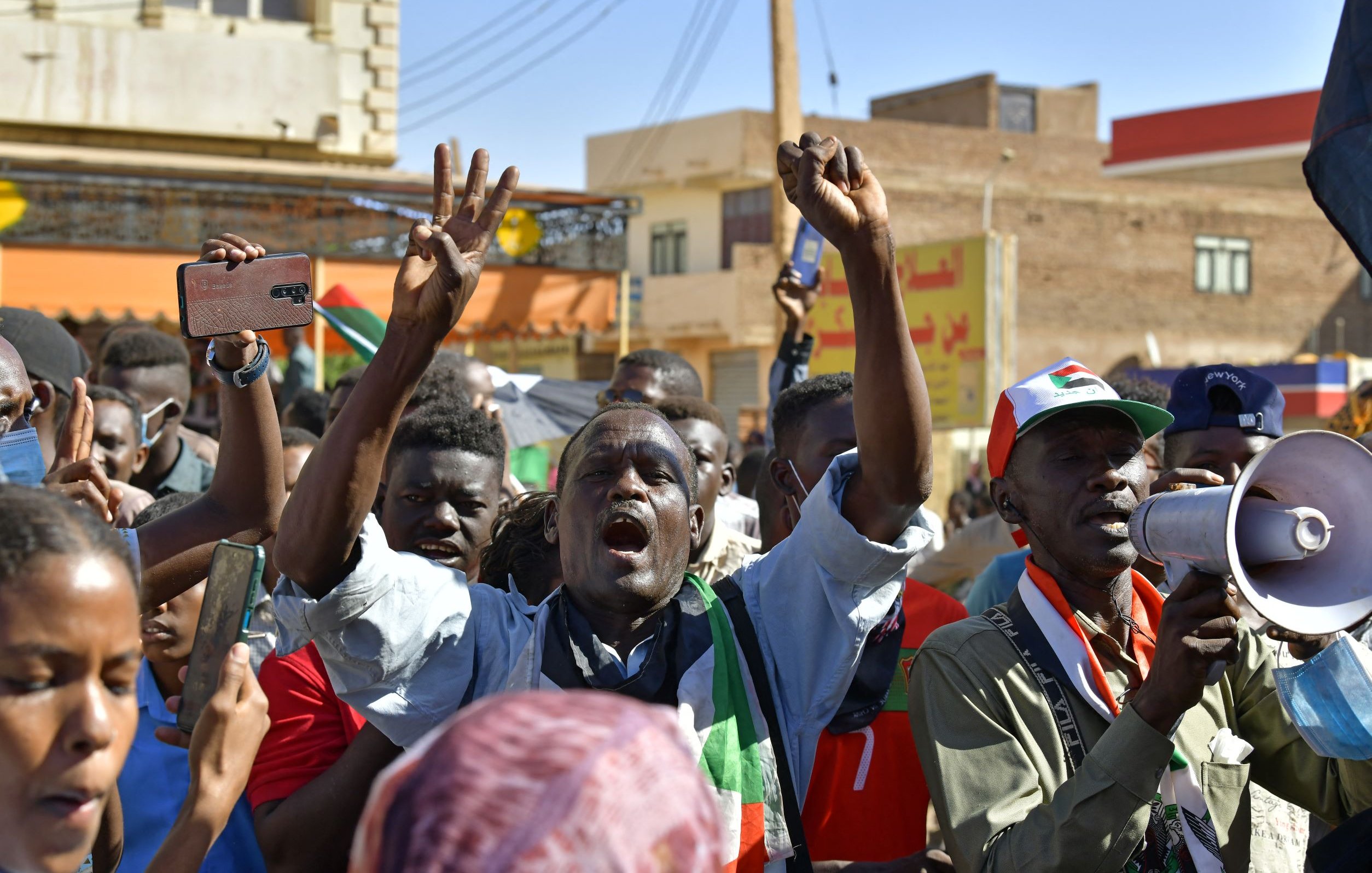 L'image montre une foule de manifestants lors d'une manifestation. Ils semblent exprimer leur colère ou leur détermination, avec des poings levés et des expressions intenses. Certaines personnes tiennent des téléphones portables, tandis qu'une autre utilise un mégaphone pour s'exprimer. En arrière-plan, on aperçoit des bâtiments et des drapeaux, ce qui donne une idée d'un contexte de protestation pour une cause déterminée. L'atmosphère semble énergique et passionnée.