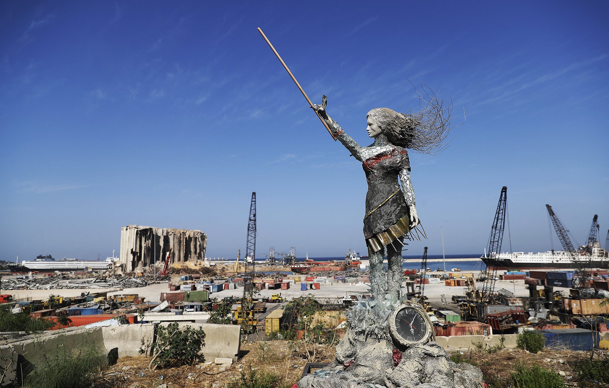 L'image présente une sculpture en métal d'une femme levant un bâton, symbolisant probablement la force et la résilience. Elle se tient sur une plateforme, surplombant un paysage industriel ou portuaire, avec des conteneurs, des grues et des bâtiments en arrière-plan. On peut également apercevoir des vestiges d'un bâtiment détruit, suggérant une scène de reconstruction ou de renouveau après une catastrophe. Le ciel est dégagé et ensoleillé, contrastant avec l'environnement chaotique du port.