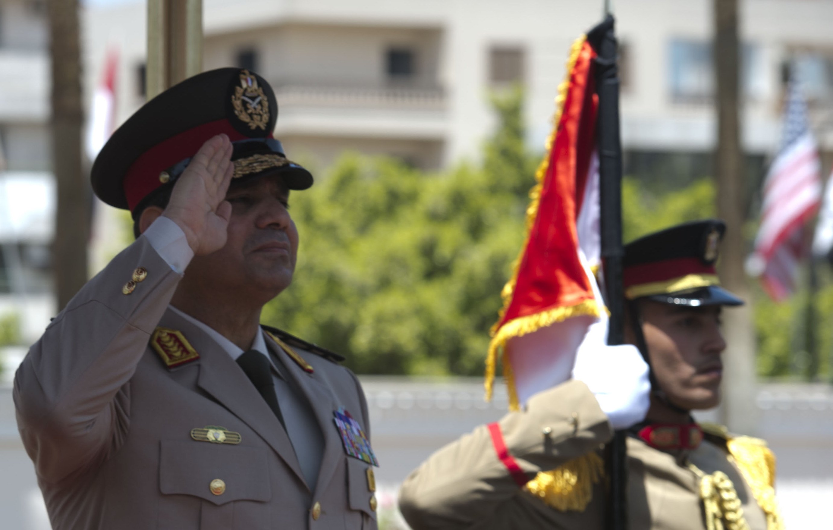 L'image représente deux officiers militaires en uniforme. L'un d'eux, probablement un haut responsable, effectue un salut militaire, tandis que l'autre maintient un drapeau. L'environnement semble être une cérémonie officielle en extérieur, avec des bâtiments en arrière-plan. Les uniformes sont ornés de médailles et de distinctions, reflétant un certain niveau de cérémonie et de formalité.