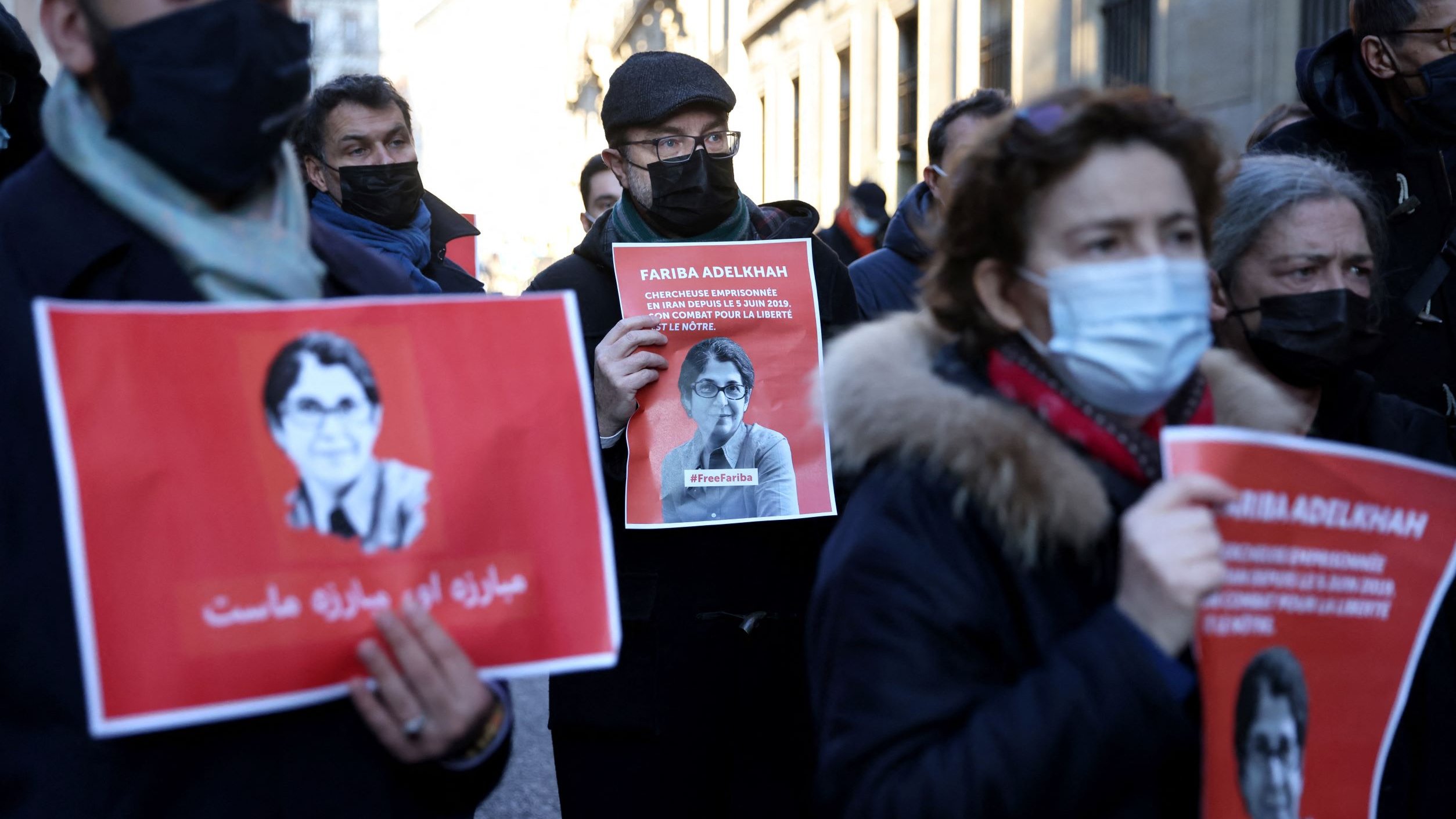 L'image montre un groupe de personnes participant à une manifestation. Elles tiennent des pancartes avec l'image et le nom d'une personne, Farida Adlénah, accompagnées de slogans. Les manifestants portent des masques, probablement en raison de préoccupations sanitaires. L'ambiance semble être solennelle et engagée, avec des visages sérieux. Les arrière-plans montrent des bâtiments qui pourraient avoir une architecture urbaine.