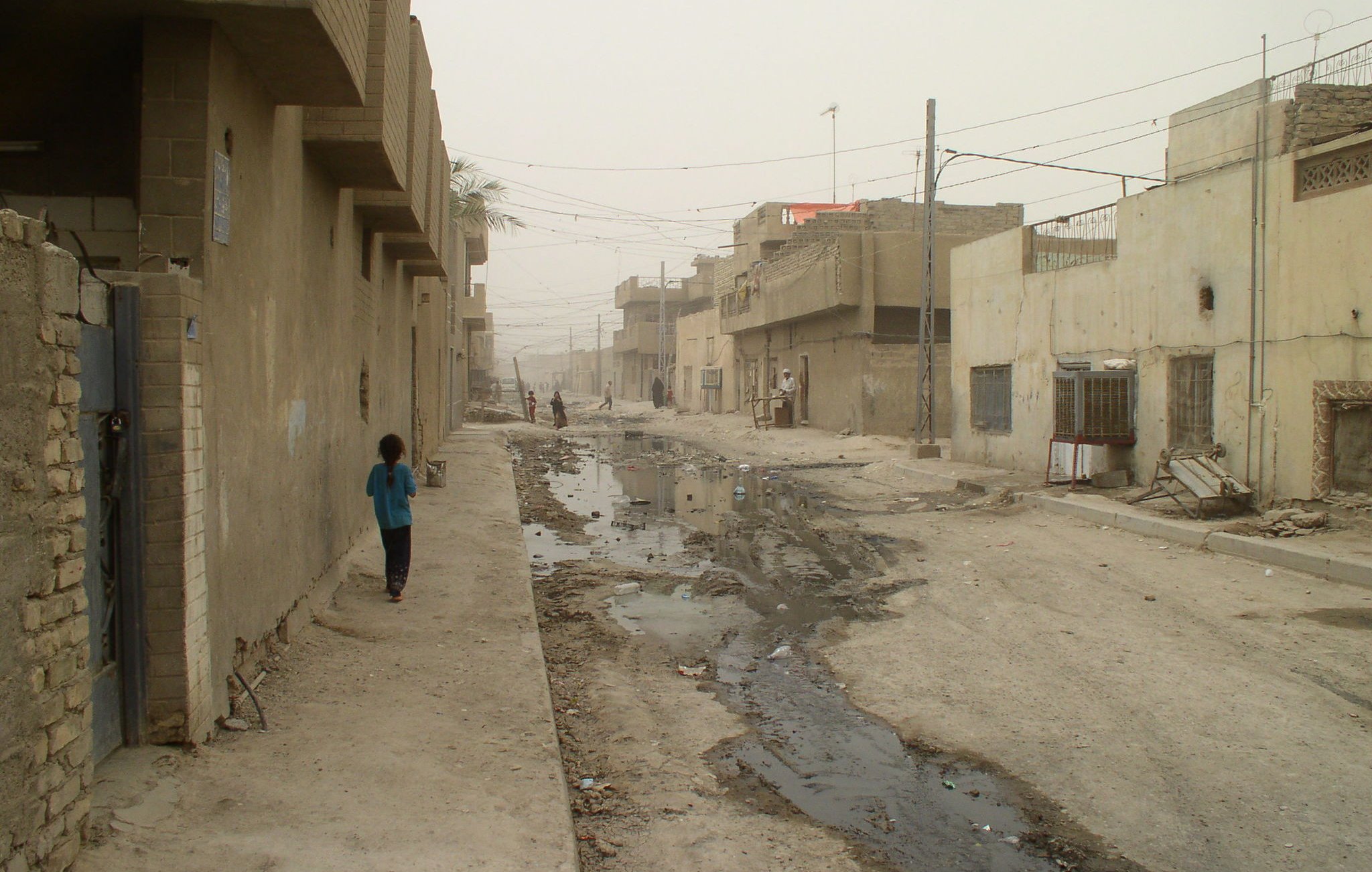 L'image montre une rue étroite et déserte dans un quartier urbain, probablement en milieu rural. Les bâtiments qui l'entourent semblent en mauvais état, avec des murs en béton et des façades peu entretenues. Le sol est inégal et présente des flaques d'eau, ce qui indique probablement une certaine négligence ou des problèmes d'infrastructure. L'atmosphère est assez grise et brumeuse, suggérant une faible visibilité, possiblement à cause de la poussière ou de la pollution. Un enfant est visible au bout de la rue, marchant seul.
