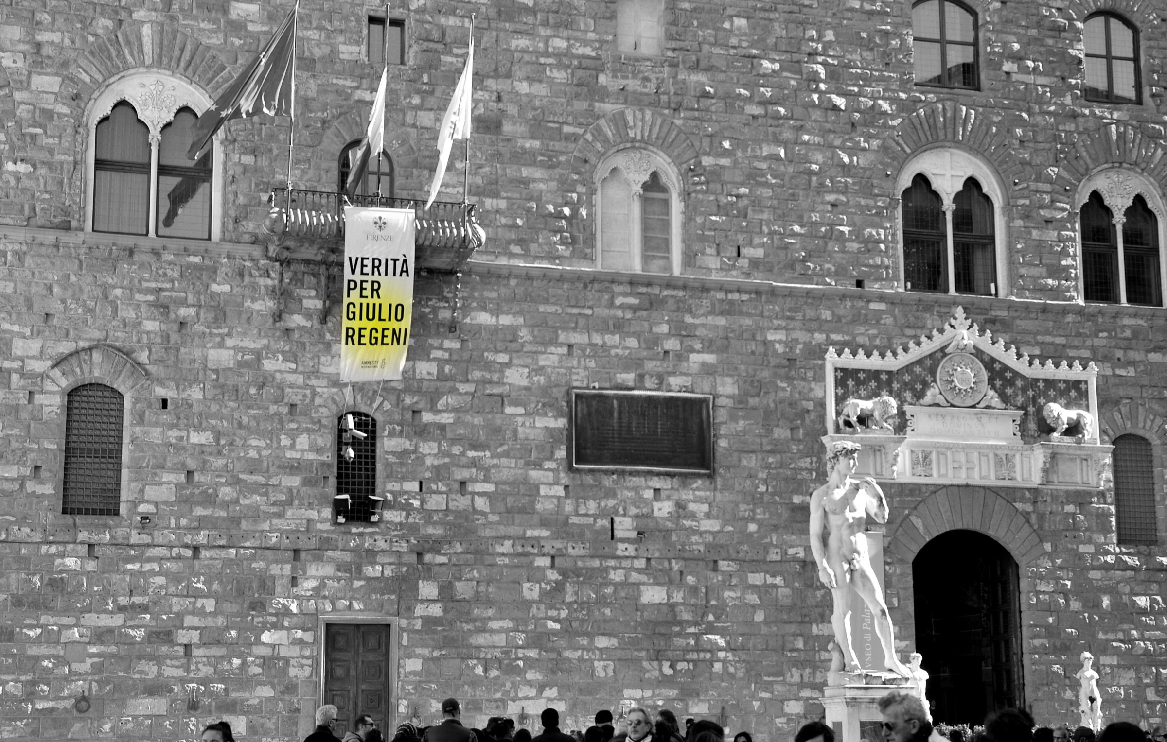 L'image montre un bâtiment historique avec une façade en pierre. Il y a une grande banderole accrochée à la façade, sur laquelle on peut lire "VERITÀ PER GIULIO REGENI", ce qui signifie "Vérité pour Giulio Regeni". En bas, on aperçoit une statue blanche, sûrement une œuvre d'art classique. Plusieurs personnes se déplacent devant le bâtiment, créant une ambiance animée. L'image est en noir et blanc, ce qui accentue le sérieux du message sur la banderole.