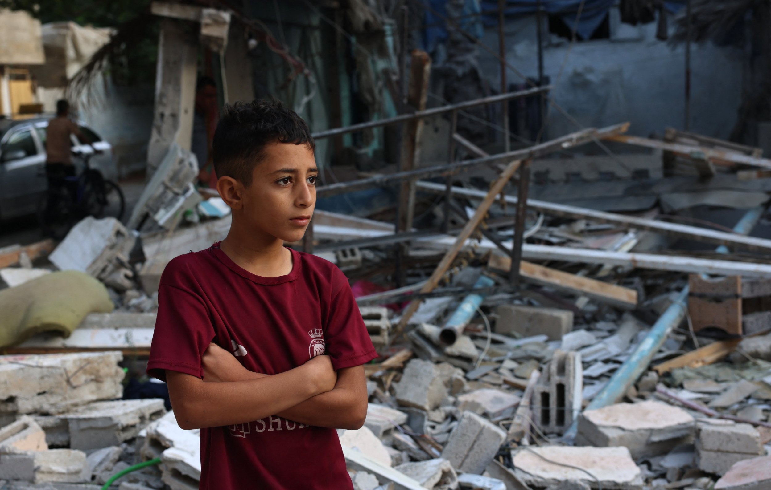 L'image montre un jeune garçon debout, les bras croisés, au milieu des débris d'un bâtiment effondré. Il semble pensif et préoccupé, exprimant une certaine gravité face à la destruction qui l'entoure. Les morceaux de béton et les matériaux épars suggèrent un environnement post-catastrophe, tandis que l'arrière-plan laisse entrevoir d'autres structures endommagées. L'atmosphère générale est sombre et témoigne d'une situation difficile.