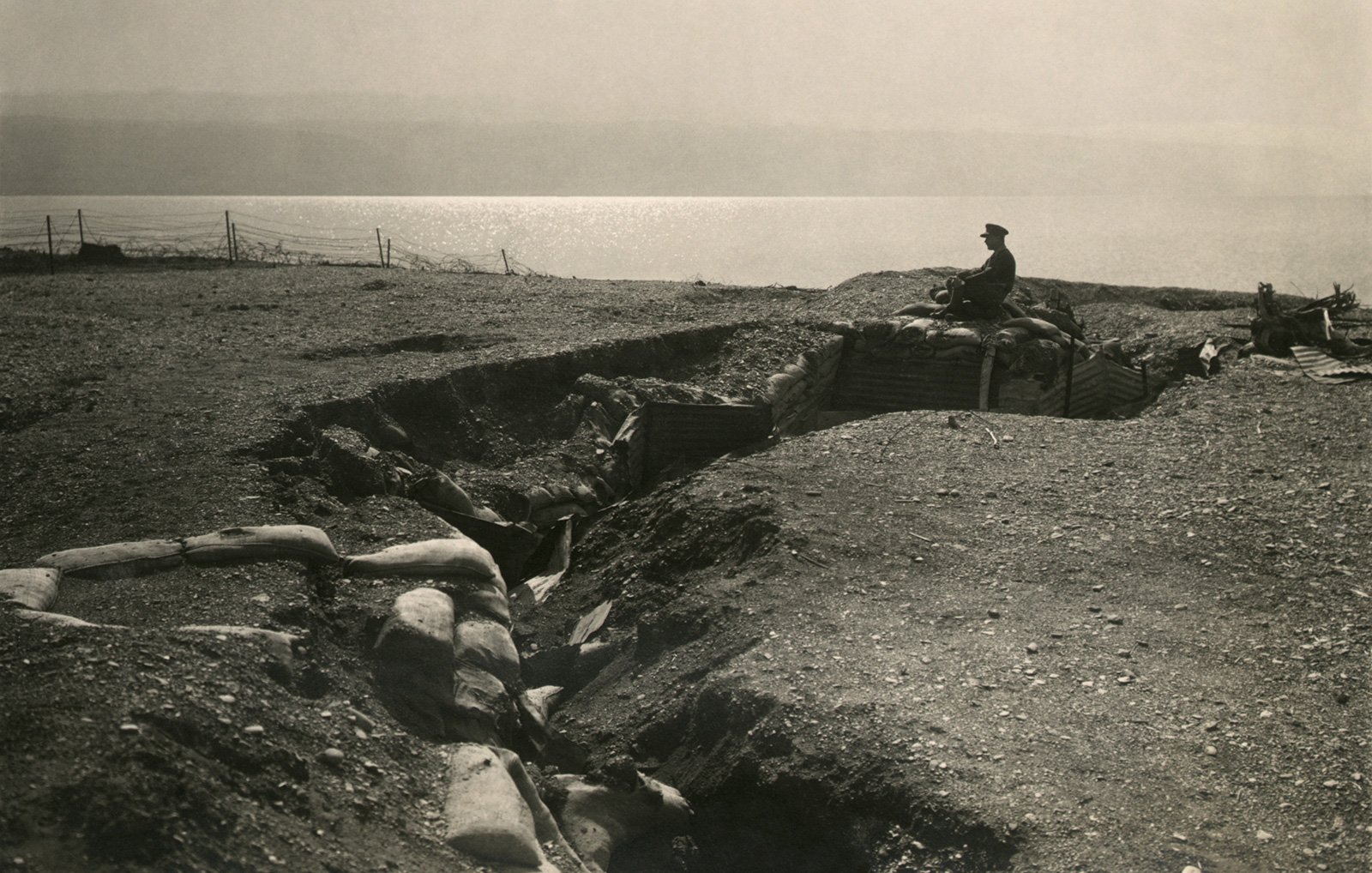 L'image représente un champ de bataille avec une tranchée. On peut voir un soldat assis sur les bords de la tranchée, qui semble surveiller les environs. Le terrain est accidenté et semé de sacs de sable, typiques des fortifications de la Première Guerre mondiale. À l'arrière-plan, on aperçoit une étendue d'eau qui pourrait être un lac ou une rivière. L'atmosphère générale est sombre et mélancolique, évoquant la dureté de la guerre.