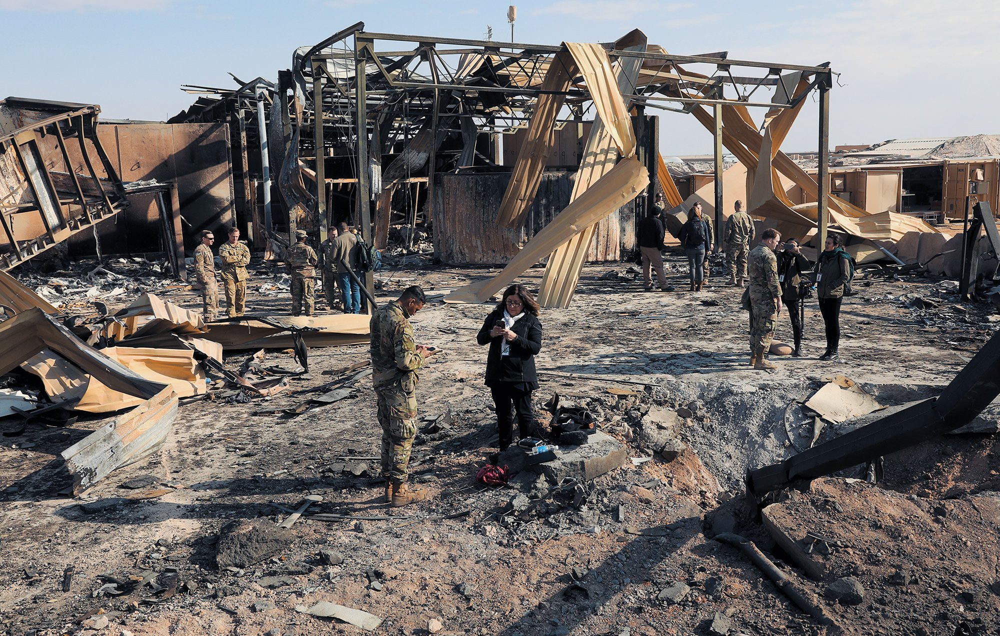 L'image montre une scène de destruction, probablement après un bombardement ou un incendie. On peut voir des débris et des structures en ruines, avec des panneaux de bois fissurés. Plusieurs militaires et civils se trouvent sur le site, certains en train de prendre des notes ou d'observer les dégâts. L'atmosphère semble grave, soulignant les conséquences d'un conflit. Le ciel est clair, contrastant avec le désastre au sol.
