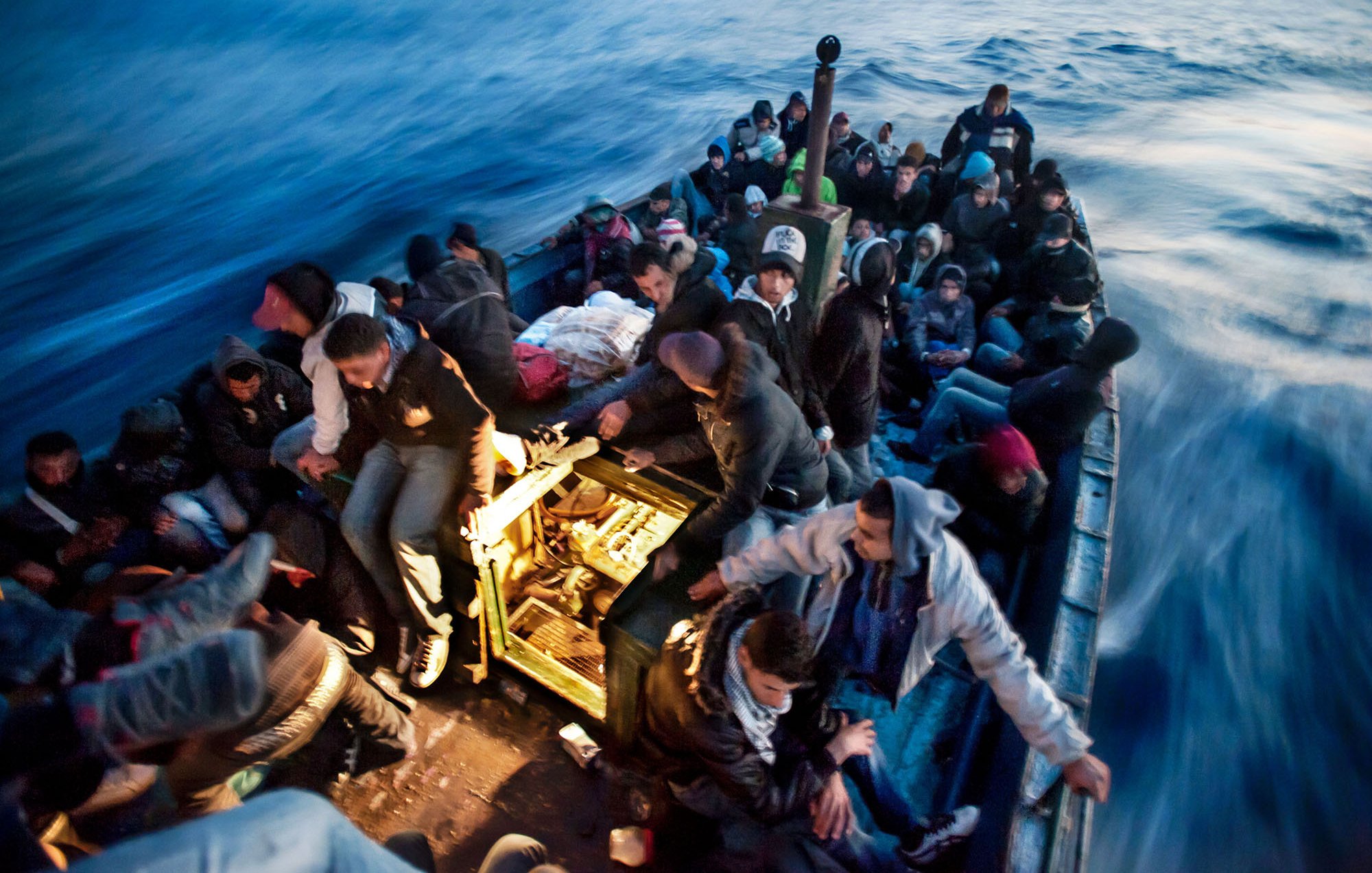 L'image montre une embarcation en mer, remplie de nombreuses personnes. Les passagers semblent être dans une situation difficile, tous étant assis ou debout, serrés les uns contre les autres. Ils portent des vêtements chauds, ce qui laisse supposer qu'ils font face à des conditions climatiques fraîches. L'eau est agitée et on peut voir des vagues, créant une atmosphère tendue et instable. La scène évoque des thèmes de migration et de recherche de refuge.