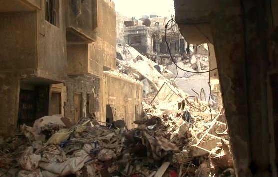 L'image montre une scène de destruction, probablement dans une zone urbaine touchée par un conflit ou un désastre. Des bâtiments sont en ruines, avec des murs effondrés et des débris éparpillés sur le sol. On peut voir des tas de gravats et de déchets, ce qui suggère que la zone a été abandonnée ou négligée. La lumière du soleil pénètre à travers les décombres, créant un contraste entre l'ombre et la lumière. L'atmosphère évoque la désolation et la tristesse.