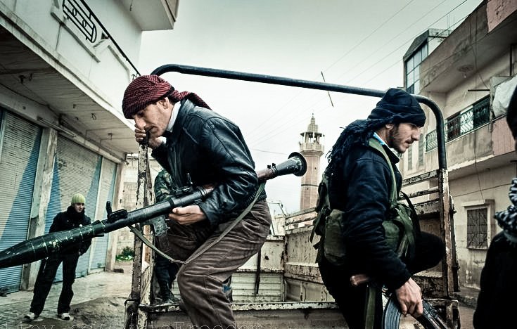L'image montre des individus armés, apparemment dans un environnement urbain. Deux hommes sont en mouvement, l'un portant une arme lourde et l'autre un fusil. Ils semblent faire partie d'un groupe en action, probablement dans un contexte de conflit. L'arrière-plan montre des bâtiments et une ambiance sombre, qui renforcent le sentiment de tension. L'atmosphère est chargée et suggère un moment urgent.