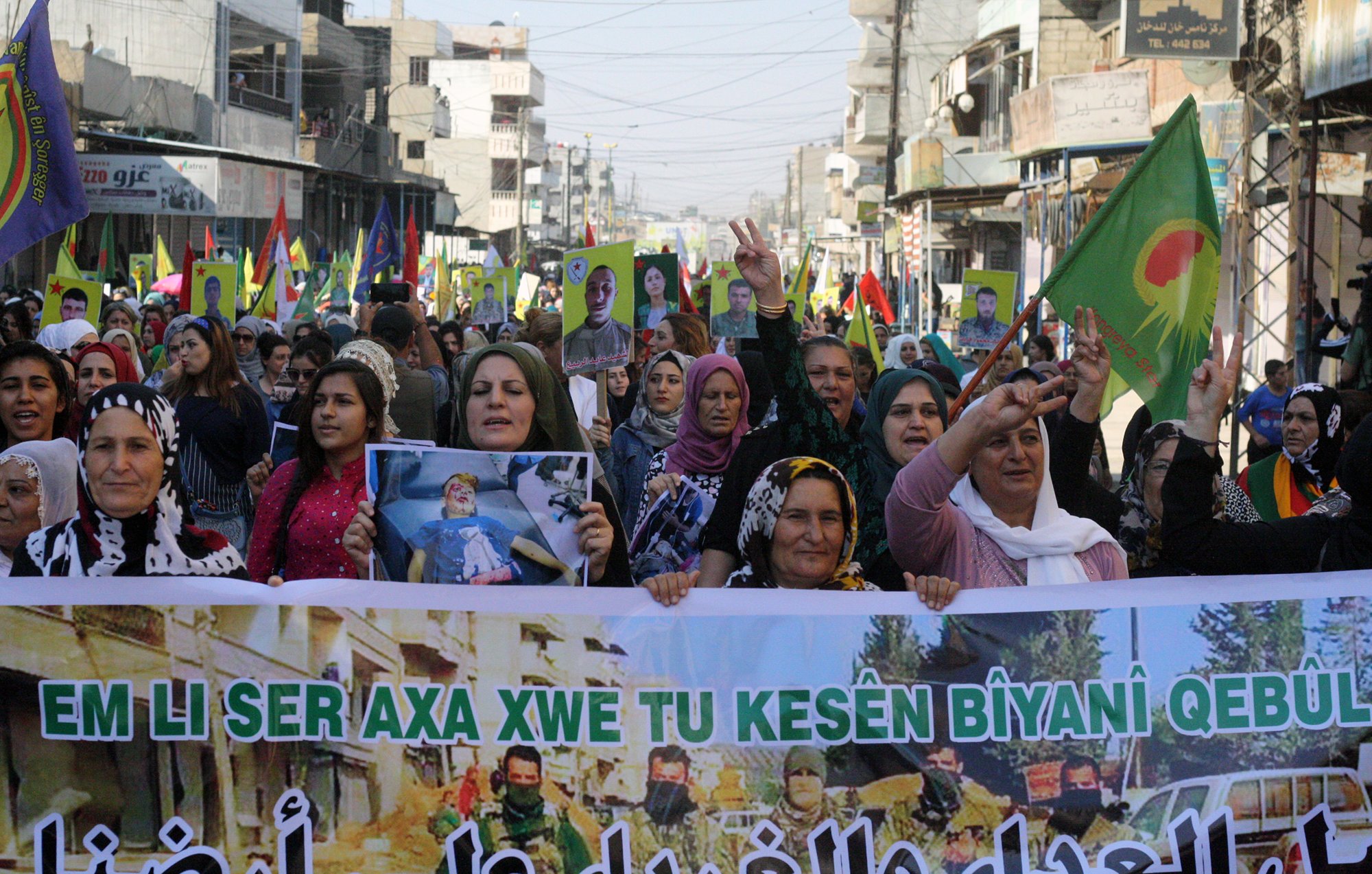 L'image montre une grande foule de personnes, principalement des femmes, participant à une manifestation. Elles portent des drapeaux colorés et affichent des pancartes avec des slogans. Les manifestants semblent exprimer des revendications et un soutien, probablement liées à des questions politiques ou sociales. L'ambiance est dynamique et déterminée, avec des signes de solidarité parmi les participants. Les rues environnantes sont visibles, ainsi que des bâtiments en arrière-plan.