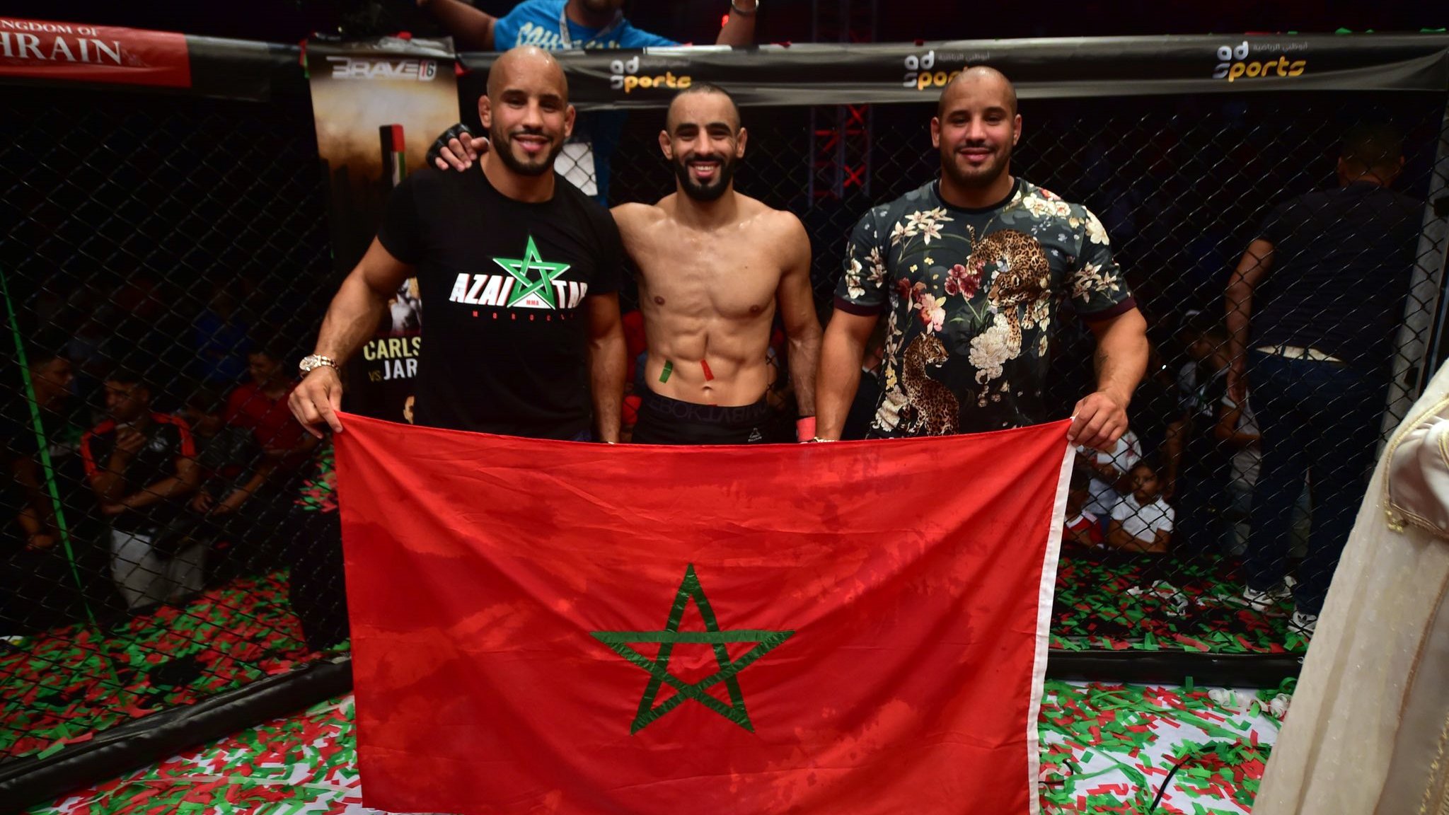 L'image montre trois hommes posant ensemble à l'intérieur d'un environnement de combat, probablement après une victoire. L'un d'eux est torse nu et affiche un large sourire, tandis que les autres sont habillés avec des t-shirts. Ils tiennent une grande drapeau marocain, mettant en avant leur fierté nationale. Le sol est couvert de confettis, ajoutant une atmosphère festive à la scène. En arrière-plan, on peut voir des spectateurs soulevant les bras en signe de célébration.