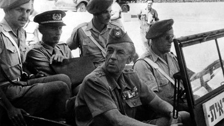 L'image montre un groupe de militaires assis dans un véhicule. Il y a plusieurs hommes en uniforme, portant des chapeaux distinctifs. Certains sont assis à l'avant, tandis que d'autres semblent être à l'arrière. L'atmosphère est solennelle, et il pourrait s'agir d'un moment lié à une opération ou une mission militaire. Le fond est flou, mais on aperçoit d'autres véhicules et un environnement urbain.