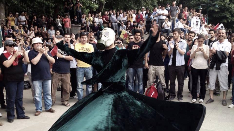 L'image montre une scène de rassemblement en plein air où une personne vêtue d'une longue robe vert foncé tourne gracieusement. Cette personne porte un masque à gaz, ce qui ajoute une dimension étrange et intrigante à la performance. Autour, une foule observe, certains prenant des photos avec leurs téléphones. L'ambiance semble à la fois festive et solennelle, avec des expressions variées parmi les spectateurs. Les arbres en arrière-plan ajoutent une touche de nature à la scène.