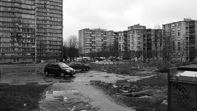 L'image montre un paysage urbain qui semble désolé. On peut voir plusieurs immeubles de grande hauteur, typiques de l'architecture des banlieues. Le sol est quelques peu inégal, avec des flaques d'eau et des débris éparpillés, ce qui donne une impression de négligence. Deux voitures sont garées sur un chemin de terre. L'environnement est sombre, accentué par le ciel nuageux, ce qui ajoute à l'atmosphère générale de tristesse et de délabrement.