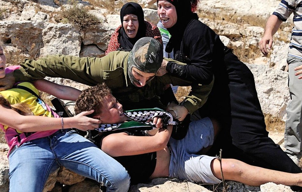 L'image montre une scène de tension où plusieurs personnes semblent être impliquées dans une confrontation. Un militaire en uniforme est en train de maîtriser un jeune homme au sol tandis que d'autres personnes, dont des femmes et des enfants, semblent réagir avec de l'indignation ou de la peur. L'environnement paraît rural, avec des rochers et une végétation sèche. L'émotion et le conflit sont palpables dans cette image.
