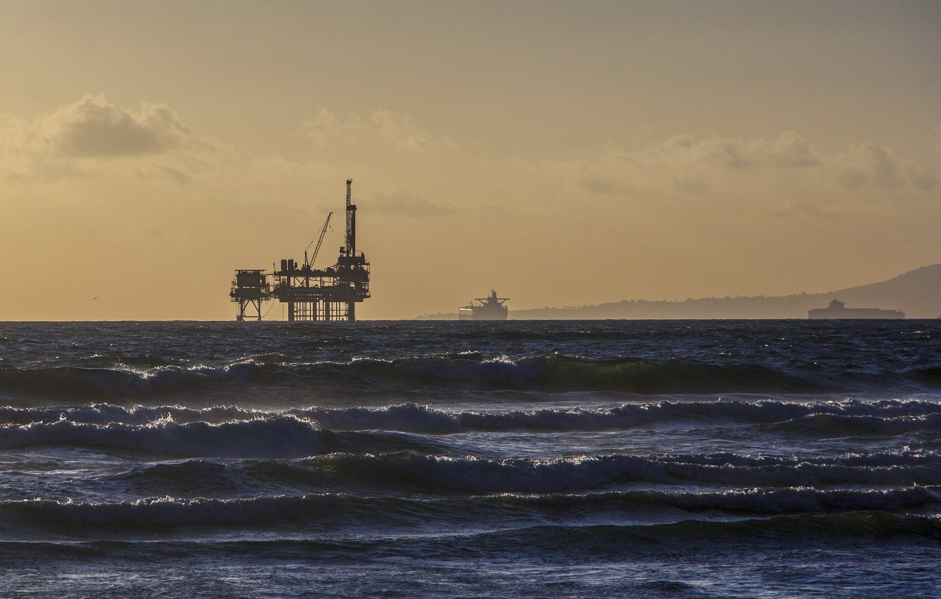 L'image montre une plateforme pétrolière située au large, se tenant fermement dans des eaux agitées. En arrière-plan, on aperçoit un navire qui navigue lentement, tandis que les vagues créent un mouvement dynamique au premier plan. La lumière du soleil se reflète sur l'eau, ajoutant une touche dorée à la scène, et le ciel présente des nuances de bleu et de beige, suggérant soit un lever soit un coucher de soleil. L'atmosphère est à la fois paisible et industrielle.