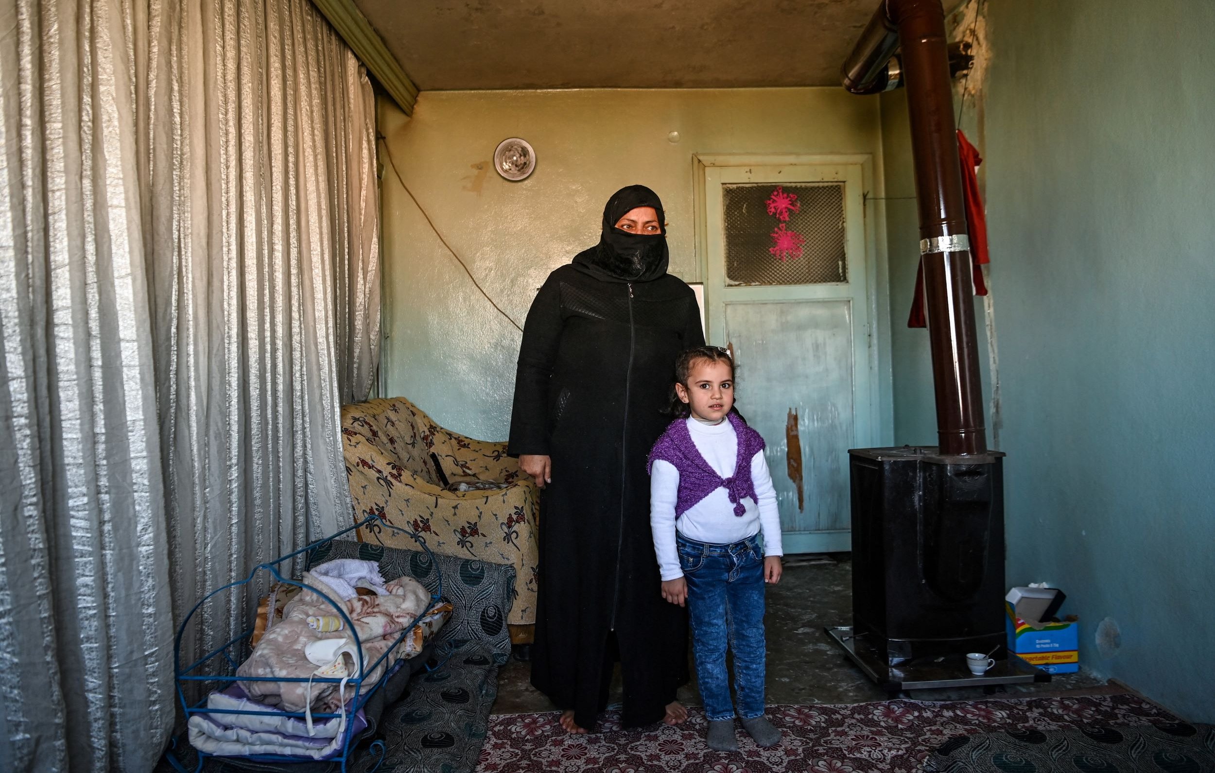 L'image montre une femme portant une tenue noire et un voile, se tenant à côté d'une petite fille. Elles sont à l'intérieur d'une pièce qui semble modeste, avec des murs verts pâles et un tapis au sol. À gauche, il y a une couchette avec un bébé endormi. Un poêle en métal est visible dans un coin de la pièce, et des rideaux en tissu léger sont tirés. L'ambiance est familiale, mais la simplicité du décor reflète une certaine précarité.
