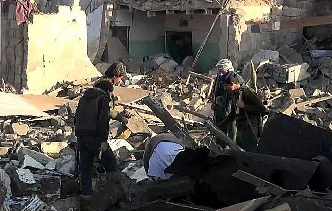 L'image montre un groupe de personnes cherchant à travers des décombres dans une zone sévèrement endommagée. Les bâtiments autour d'eux semblent en ruines, avec des murs effondrés et de nombreux débris éparpillés sur le sol. Les individus semblent concentrés et préoccupés, cherchant possiblement des survivants ou des biens perdus dans les débris. L'environnement est chaotique et témoigne d'une grande destruction.