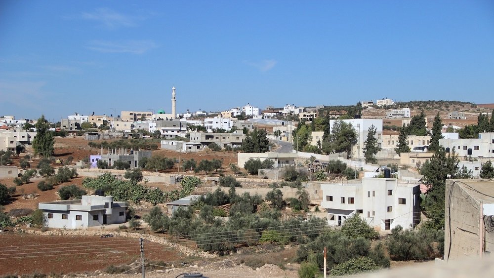 L'image montre un paysage urbain de type rural, avec des maisons de couleur claire disséminées sur une colline. On peut apercevoir des arbres et des champs, ainsi qu'un minaret se dressant dans le fond, ce qui pourrait indiquer la présence d'une mosquée. Le ciel est dégagé et d'un bleu vif, ce qui donne à la scène une atmosphère ensoleillée. Les infrastructures semblent simples et typiques des zones périphériques.