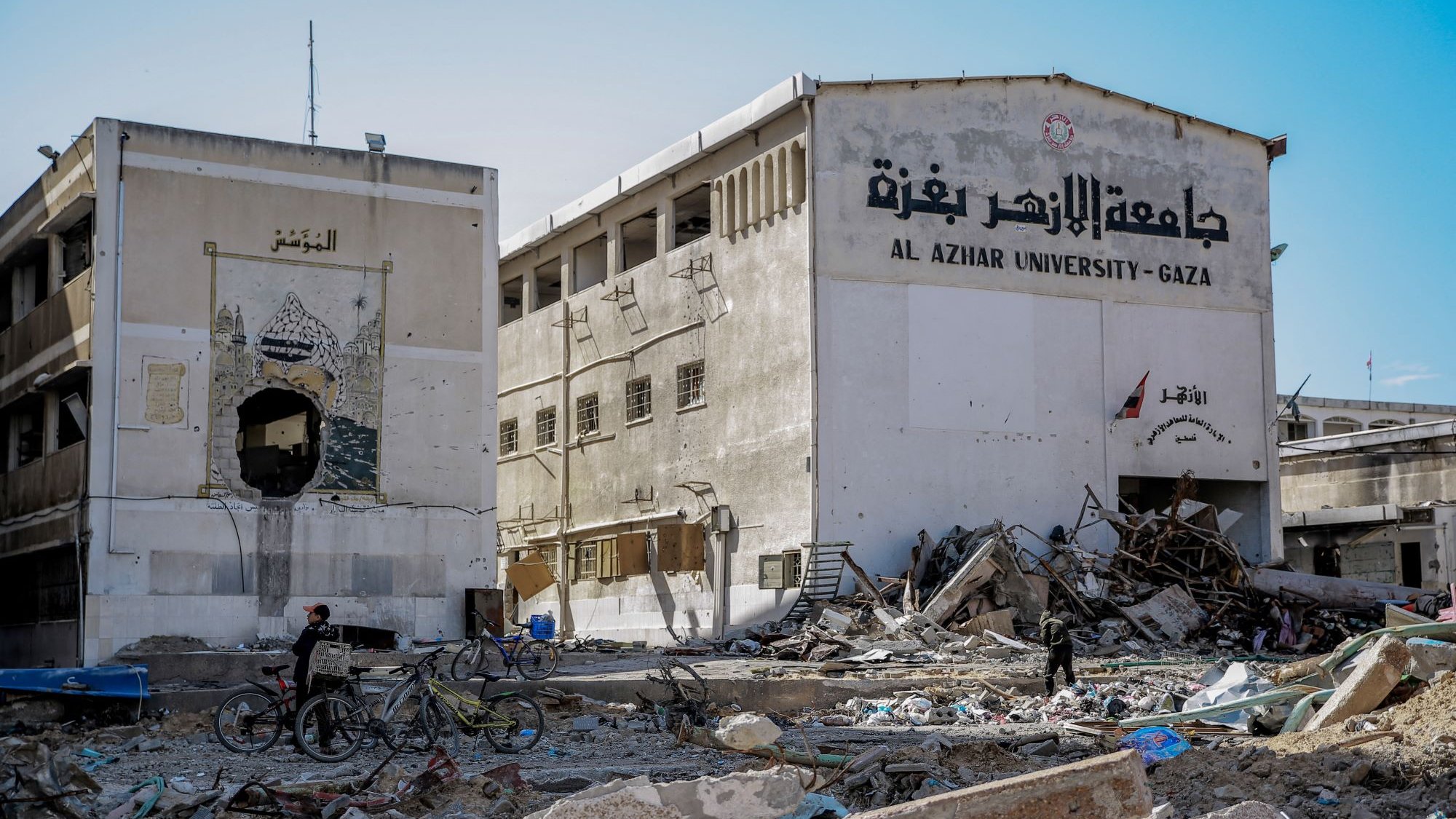 L'image montre un paysage de destruction, probablement à Gaza. On aperçoit un bâtiment, vraisemblablement l'Université Al-Azhar, dont la façade est endommagée. Des débris et des gravats jonchent le sol, indiquant des destructions significatives. Deux personnes à vélo se déplacent parmi les débris, ce qui souligne la difficulté de la situation. Le mur du bâtiment est partiellement effondré, et des inscriptions en arabe sont visibles. L'atmosphère de l'image est sombre, révélant les conséquences d'un conflit.