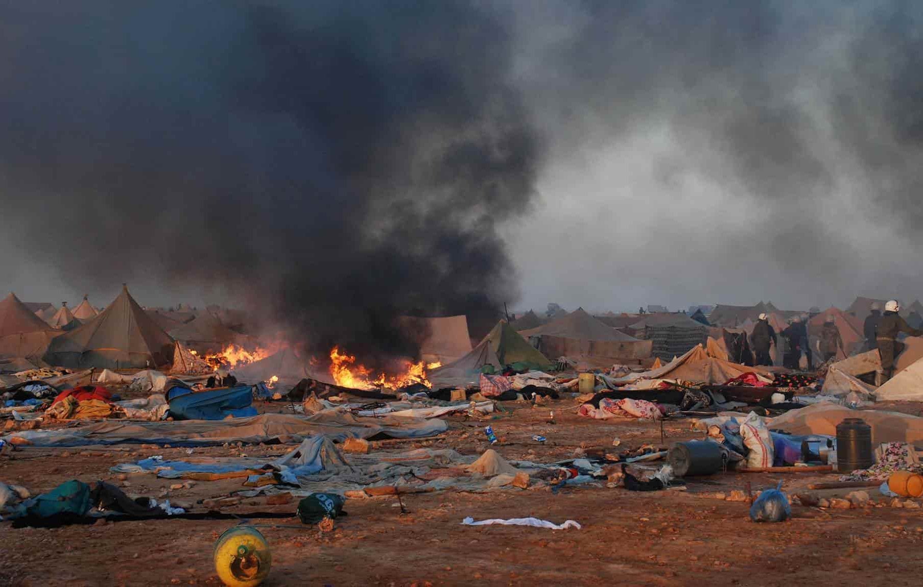 L'image montre un camp de tentes dans un environnement chaotique. On peut voir de la fumée noire s'élever dans le ciel, accompagnée de flammes, indiquant un incendie. Le sol est encombré de débris et de couvertures abandonnées, ce qui suggère une situation difficile. En arrière-plan, des personnes sont visibles, probablement des secouristes ou des autorités, tentant de gérer la situation. L'atmosphère est sombre et perturbée, reflétant une scène de crise ou de catastrophe.